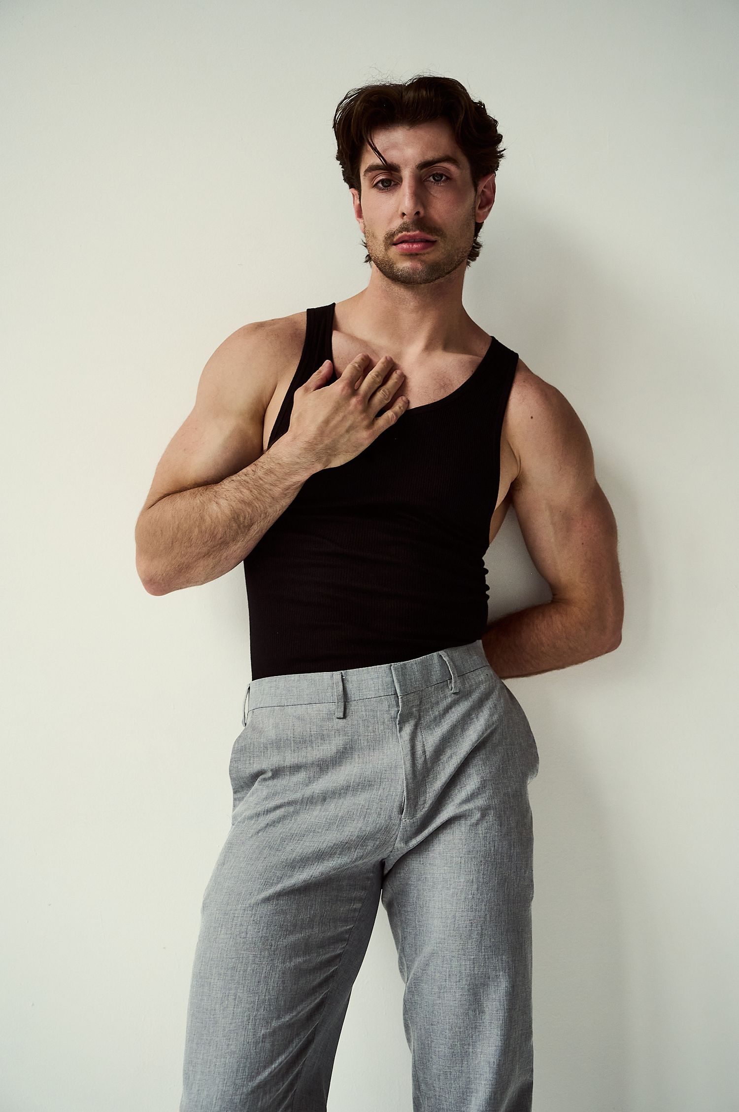 Muscular man in black tank top and gray trousers posing against white wall with hand on chest.
