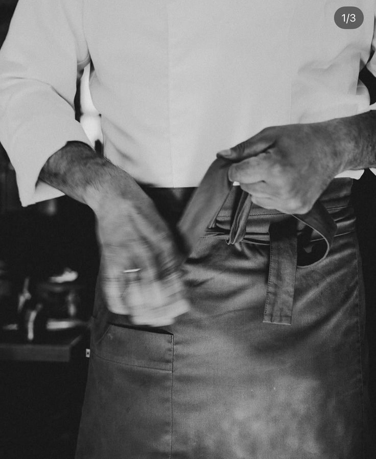 Black and white photo of a chef in white uniform adjusting a dark leather apron around their waist.