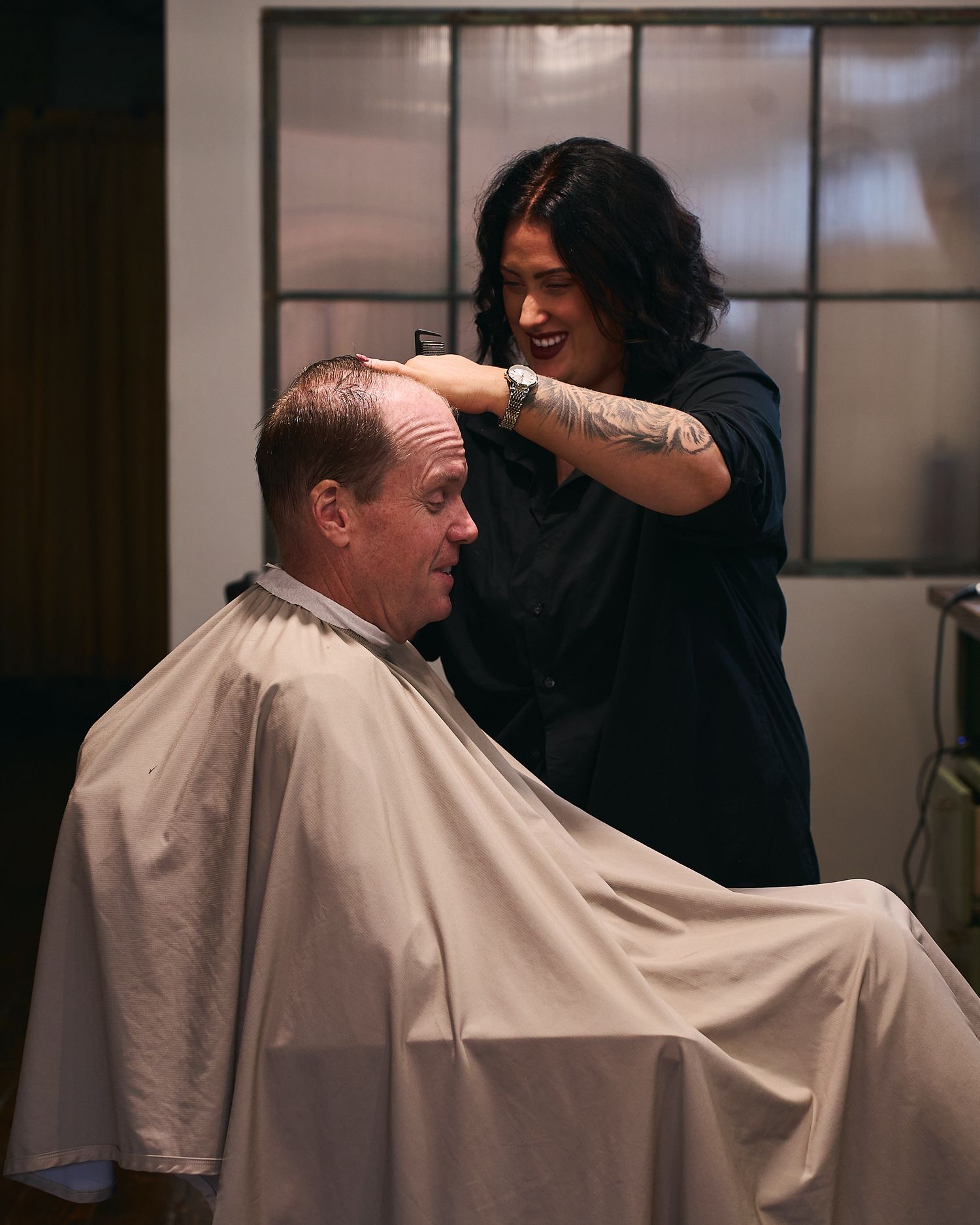 Female barber with tattoos smiling while cutting a mans hair in a modern barbershop.