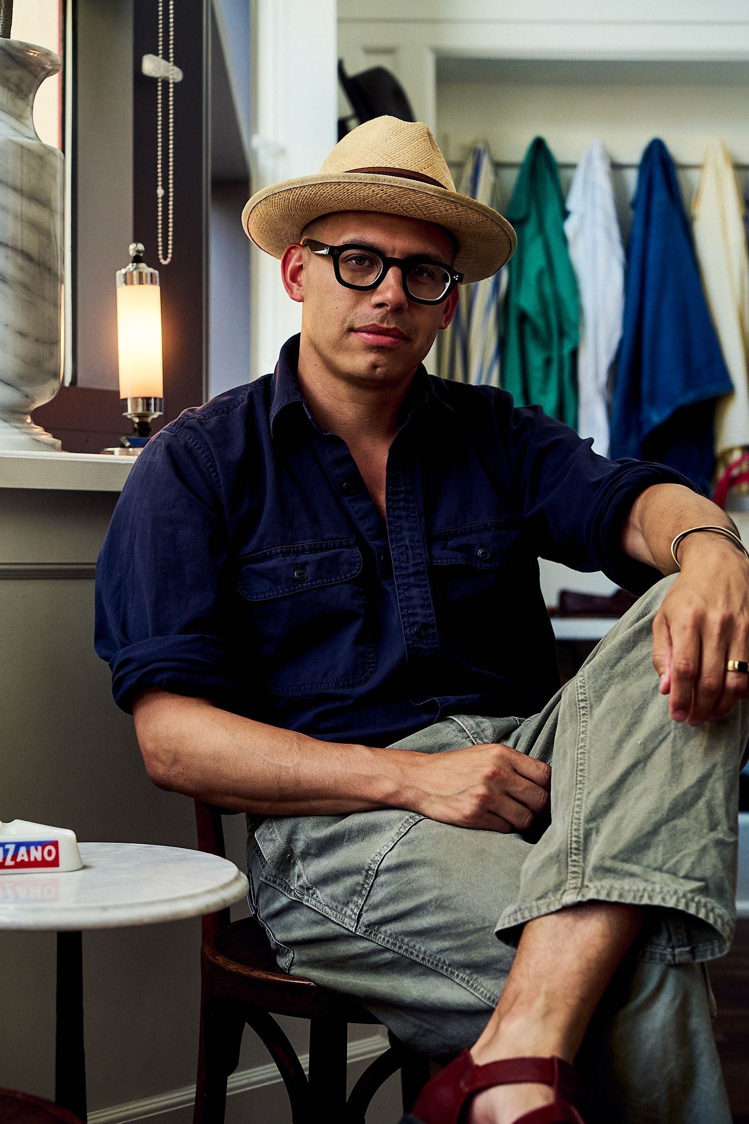 Stylish man wearing a straw hat and glasses sitting in a clothing boutique with colorful garments in background.
