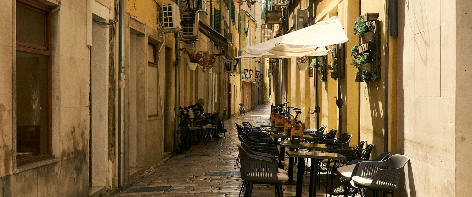 Narrow cobblestone alley with outdoor cafe chairs and tables beneath a white umbrella in an old European town.