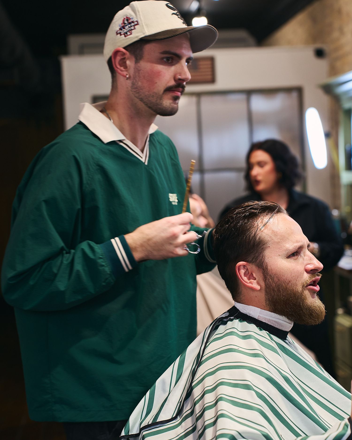 Barber in green jacket and cap cutting a clients hair with scissors in a busy barbershop.