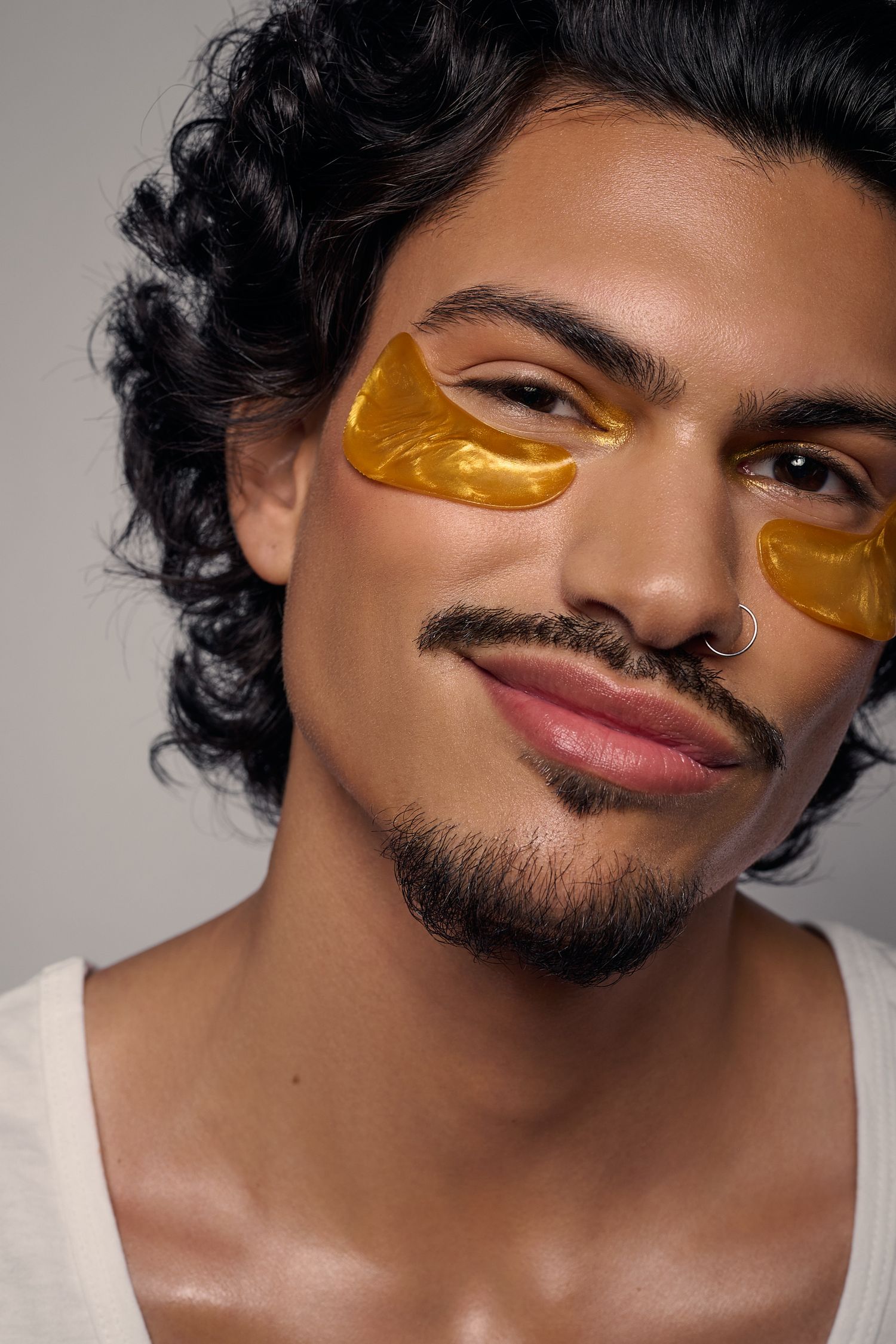 Portrait of a man with curly hair and a nose ring wearing golden under-eye patches on his face.