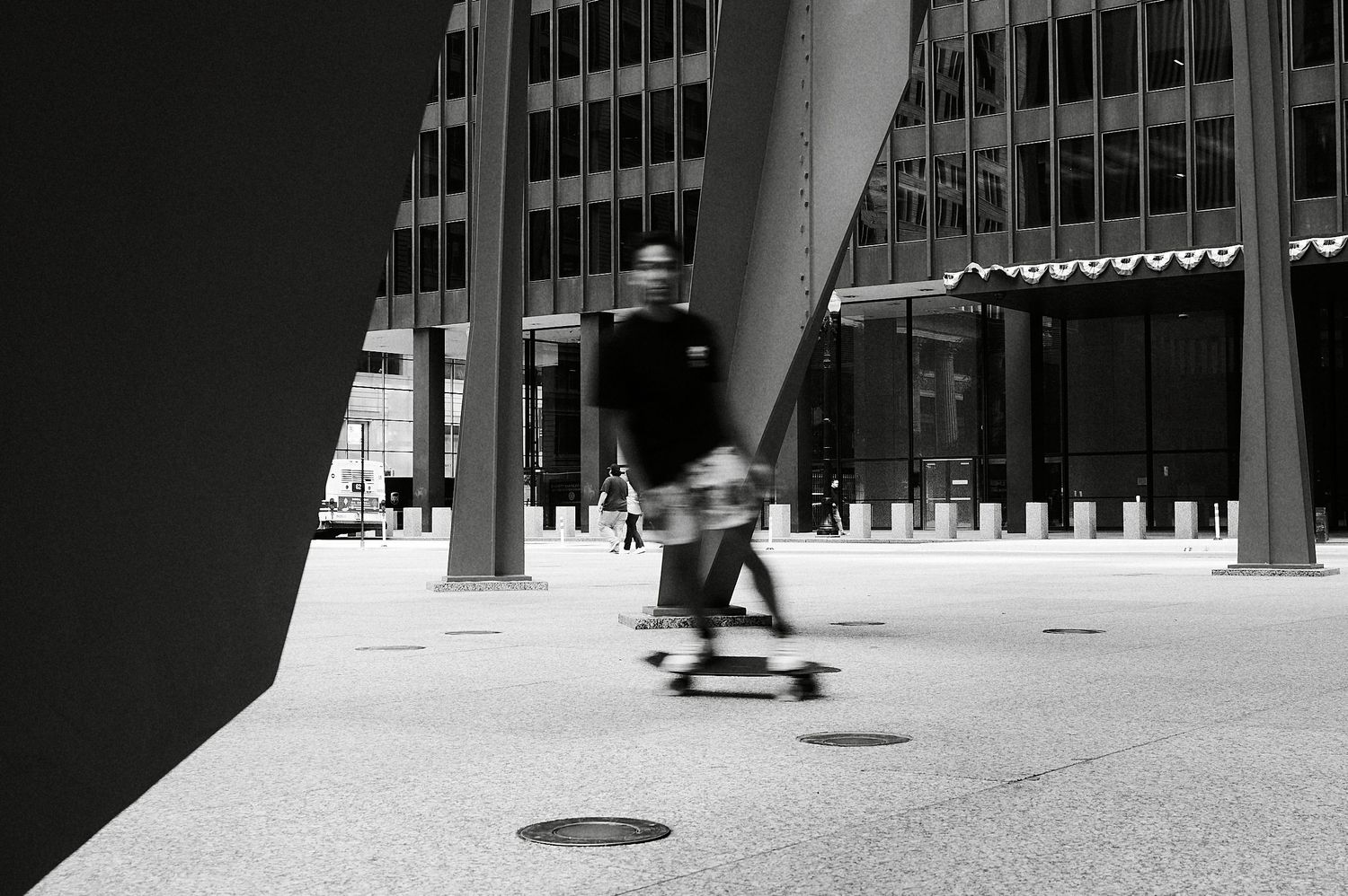 Black and white photo of a blurred man skateboarding past modern office buildings on an urban plaza.