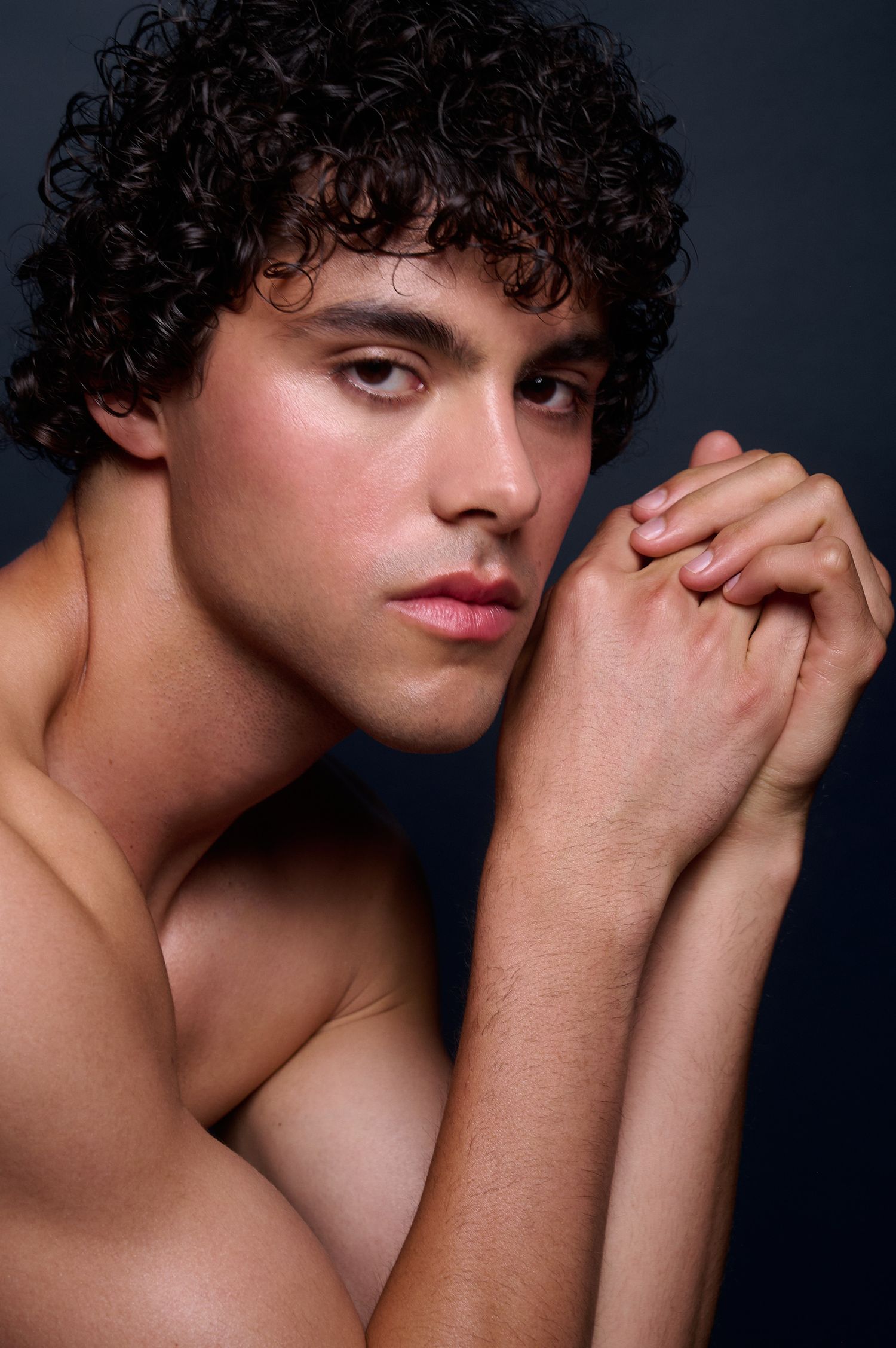 Young man with curly dark hair clasping hands near face against dark background.