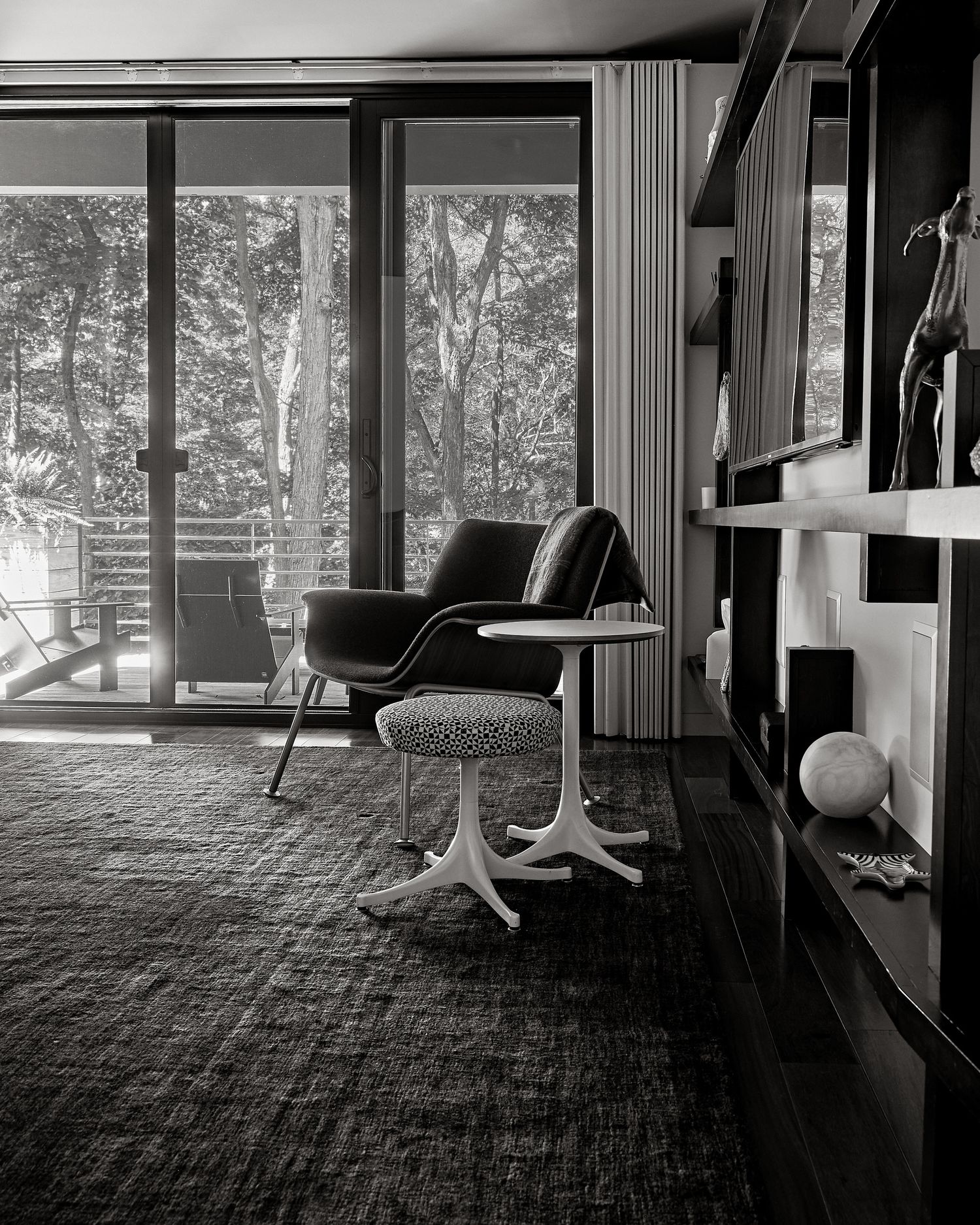 Black and white mid-century modern interior with lounge chair, tulip side table, and floor-to-ceiling windows overlooking trees.