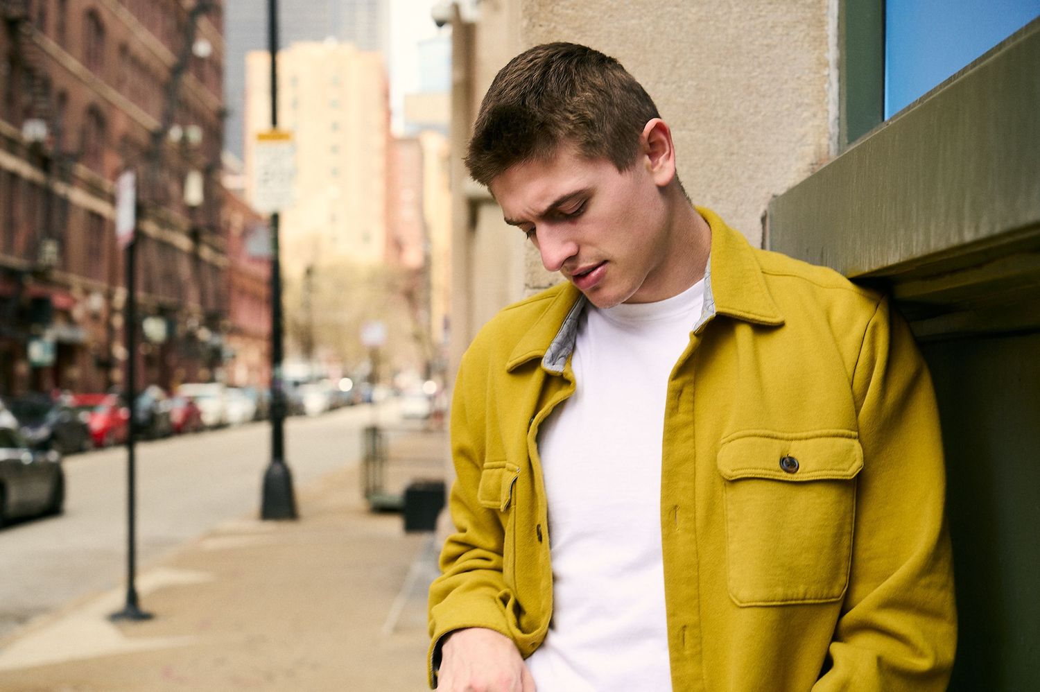 Young man in yellow jacket leaning against building on urban street, looking down thoughtfully.