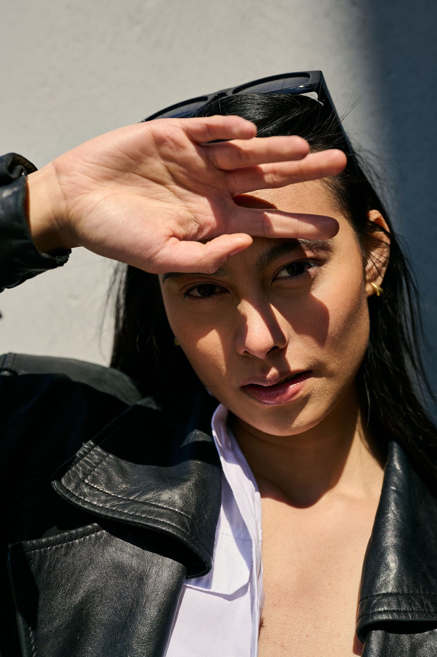 Woman in leather jacket shielding eyes from sun with hand, sunglasses on head, against white wall.