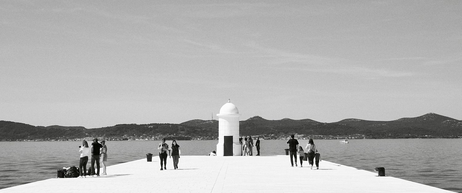People strolling on a long white pier extending into calm sea waters with mountains in the background.