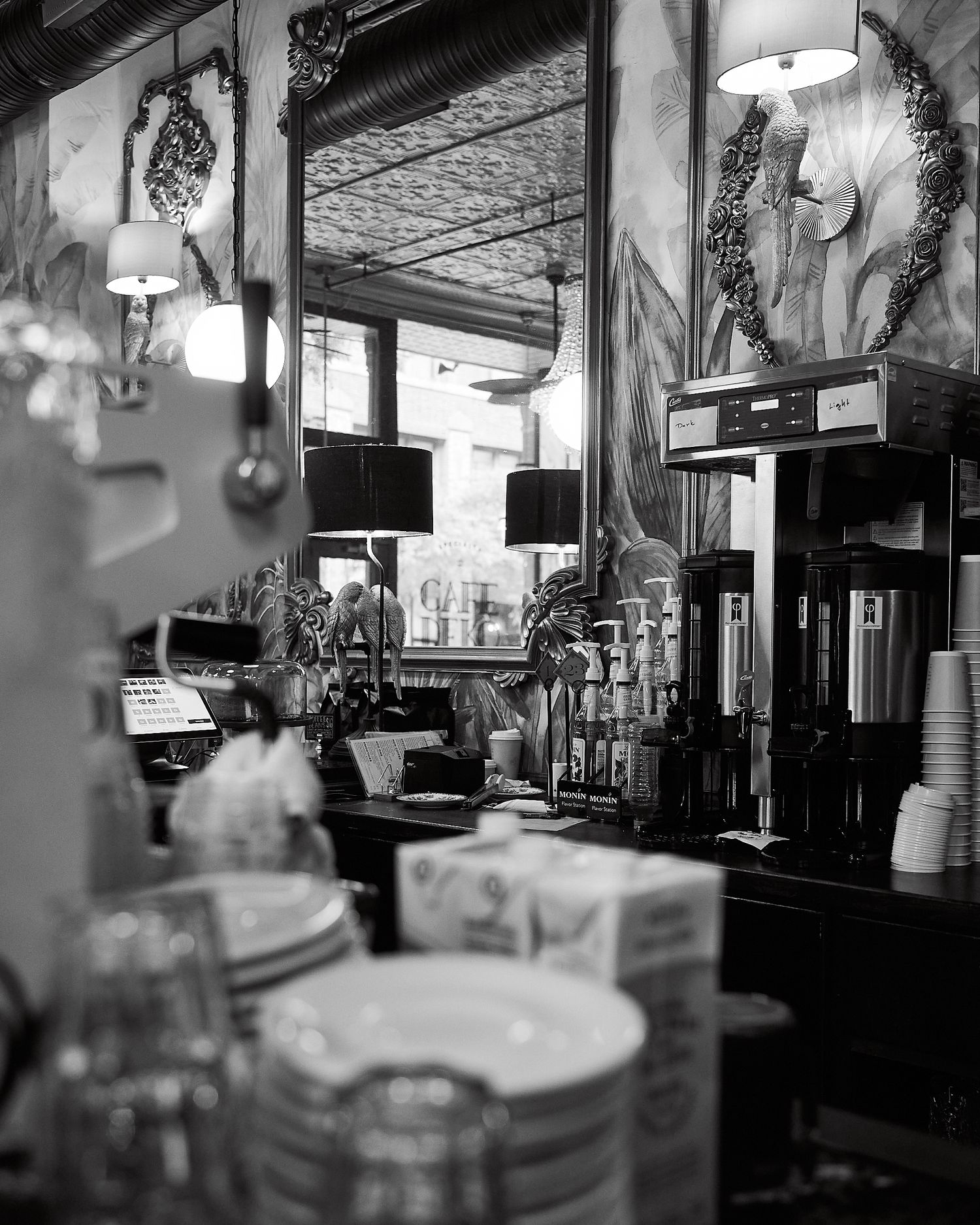 Black and white photo of a vintage cafe interior with stacked dishes, coffee equipment, and ornate mirrors on the walls.