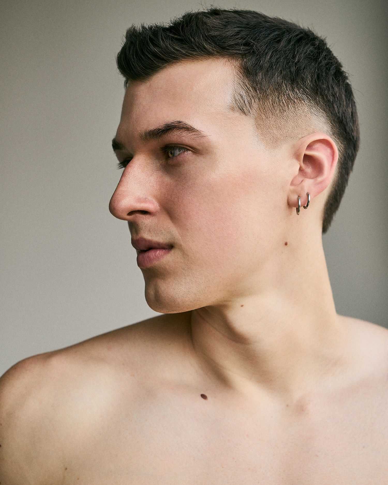 Young man with short dark hair, small hoop earring, looking sideways against neutral background.