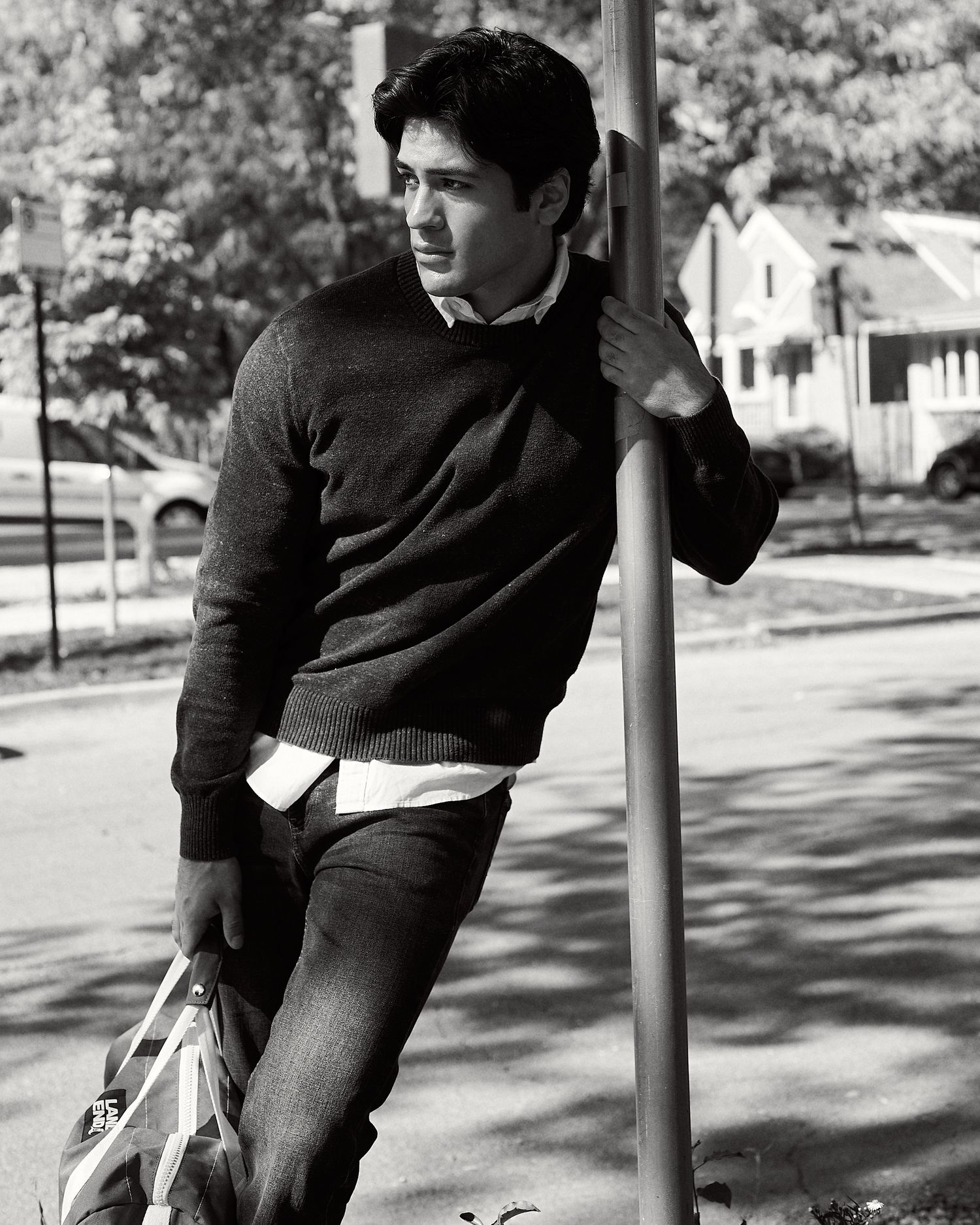 Black and white photo of a young man leaning against a pole outdoors, wearing a sweater and jeans.