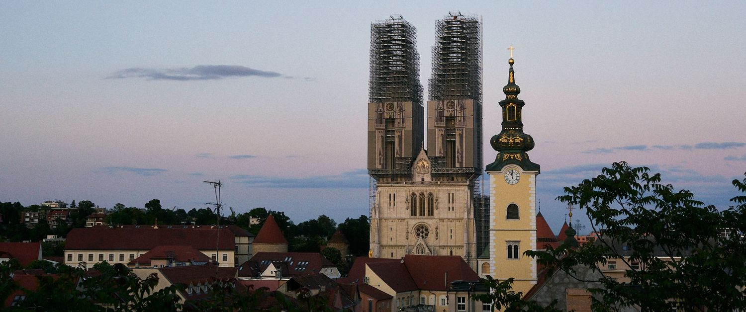Zagreb Cathedral with twin towers under scaffolding at dusk, with St. Mary Church visible against a pastel sky.