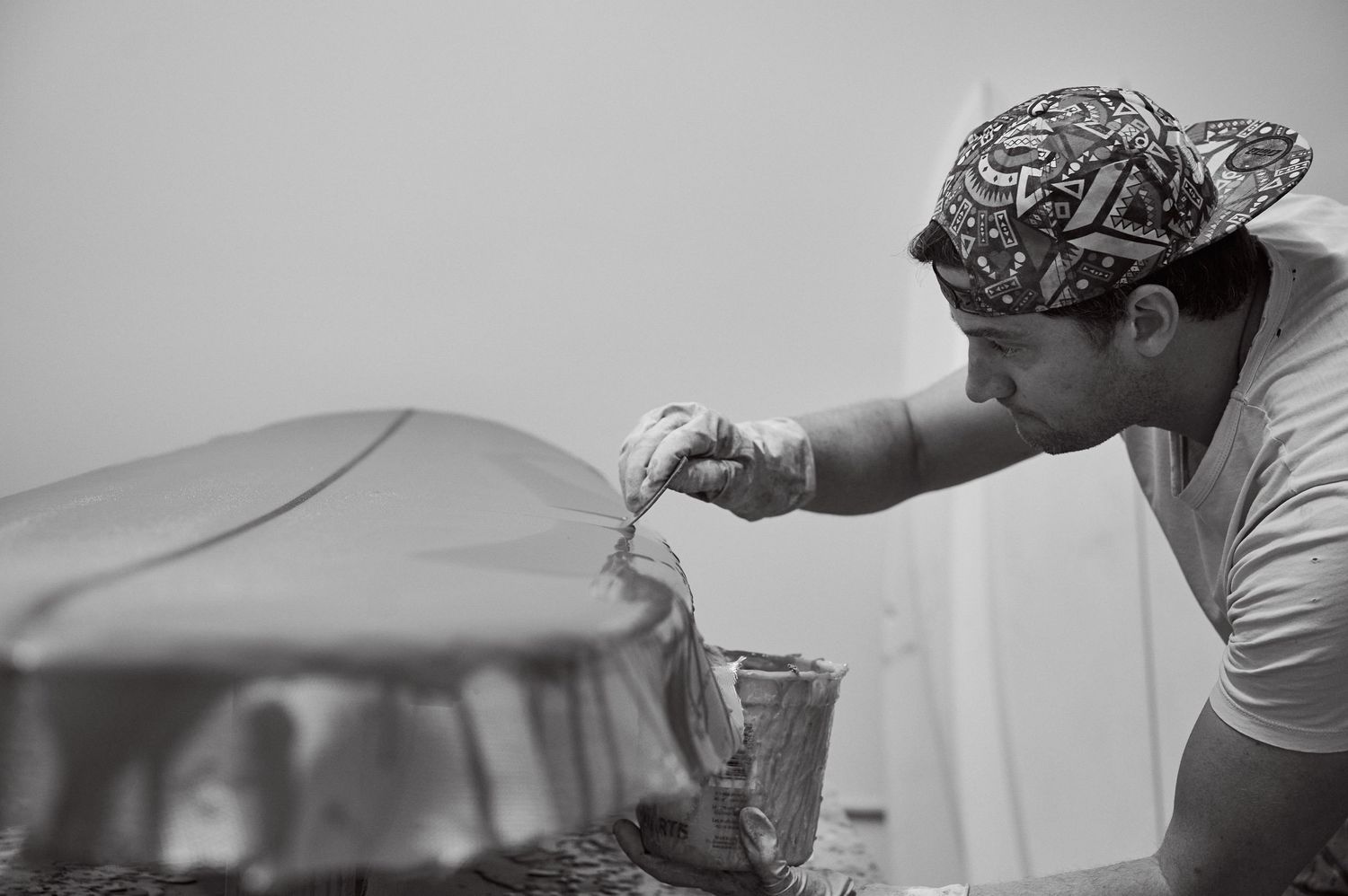 Craftsman shaping surfboard in workshop wearing patterned bandana, black and white photo.