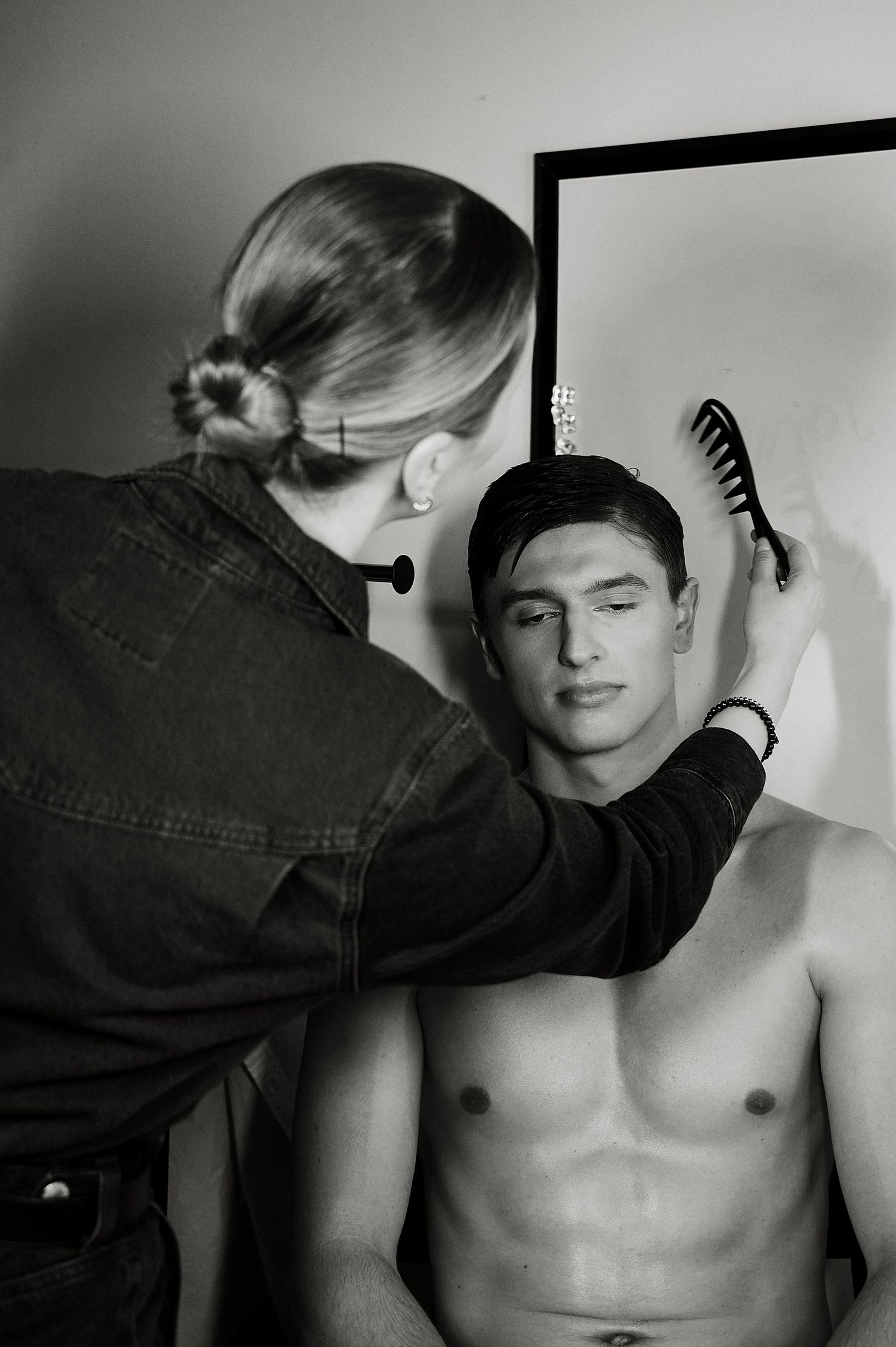 Black and white behind-the-scenes photo of a stylist combing a shirtless male model's hair.