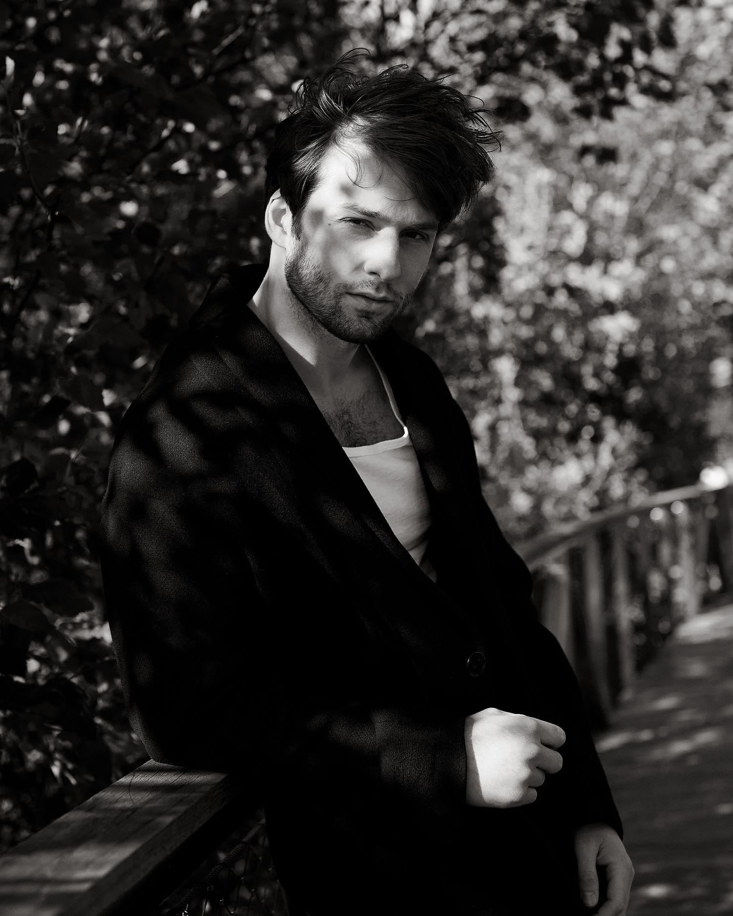 Black and white outdoor portrait of a man in a dark coat leaning against a railing among trees.