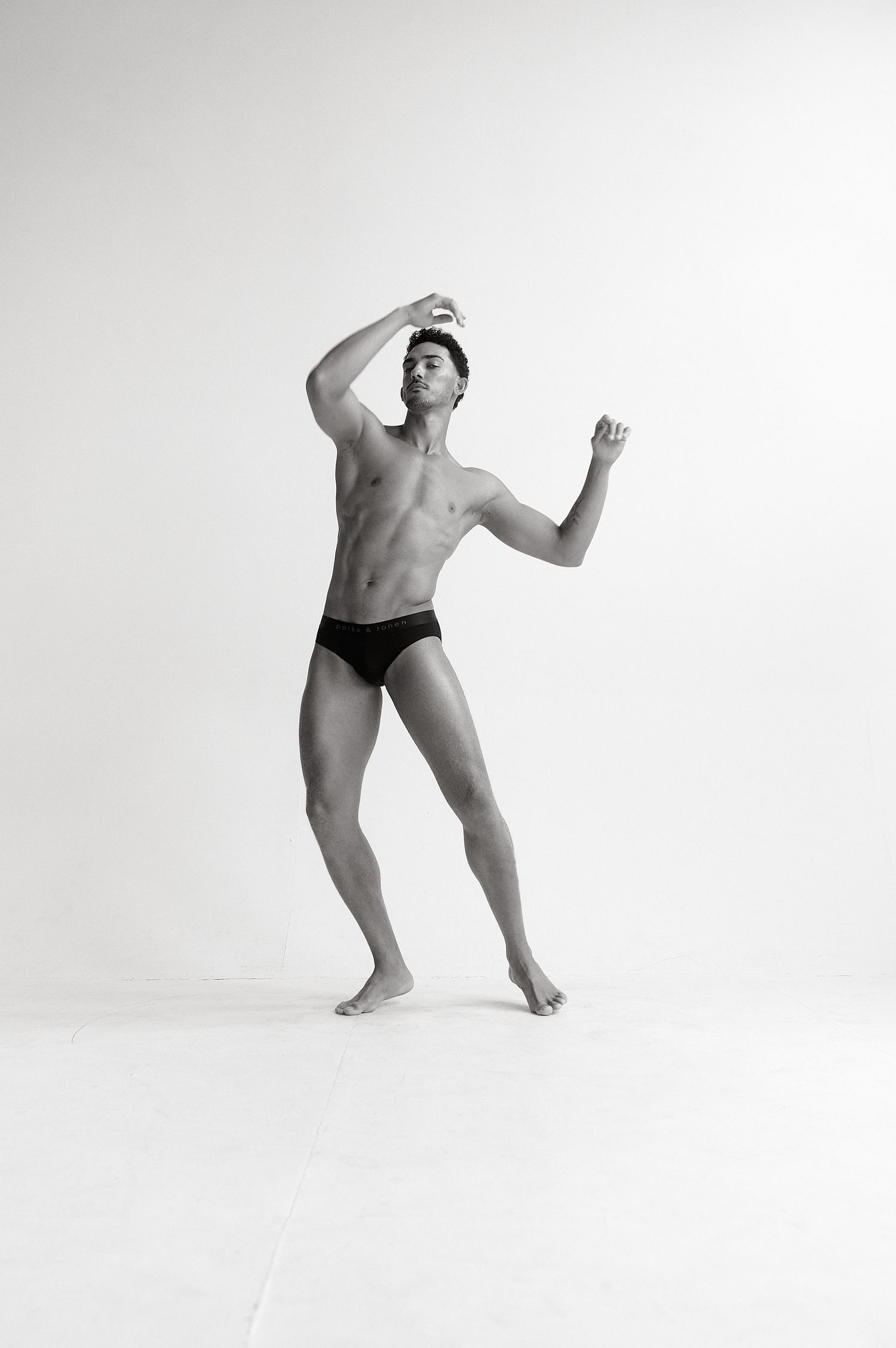 Muscular male dancer posing shirtless in black briefs against white background in black and white photo.