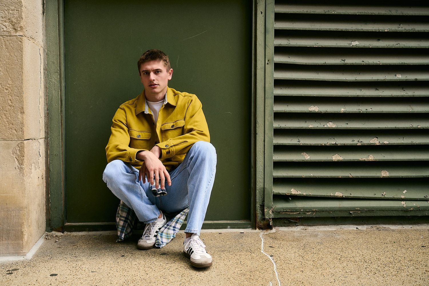 Young man in yellow jacket and jeans sitting casually against a green wall outdoors.