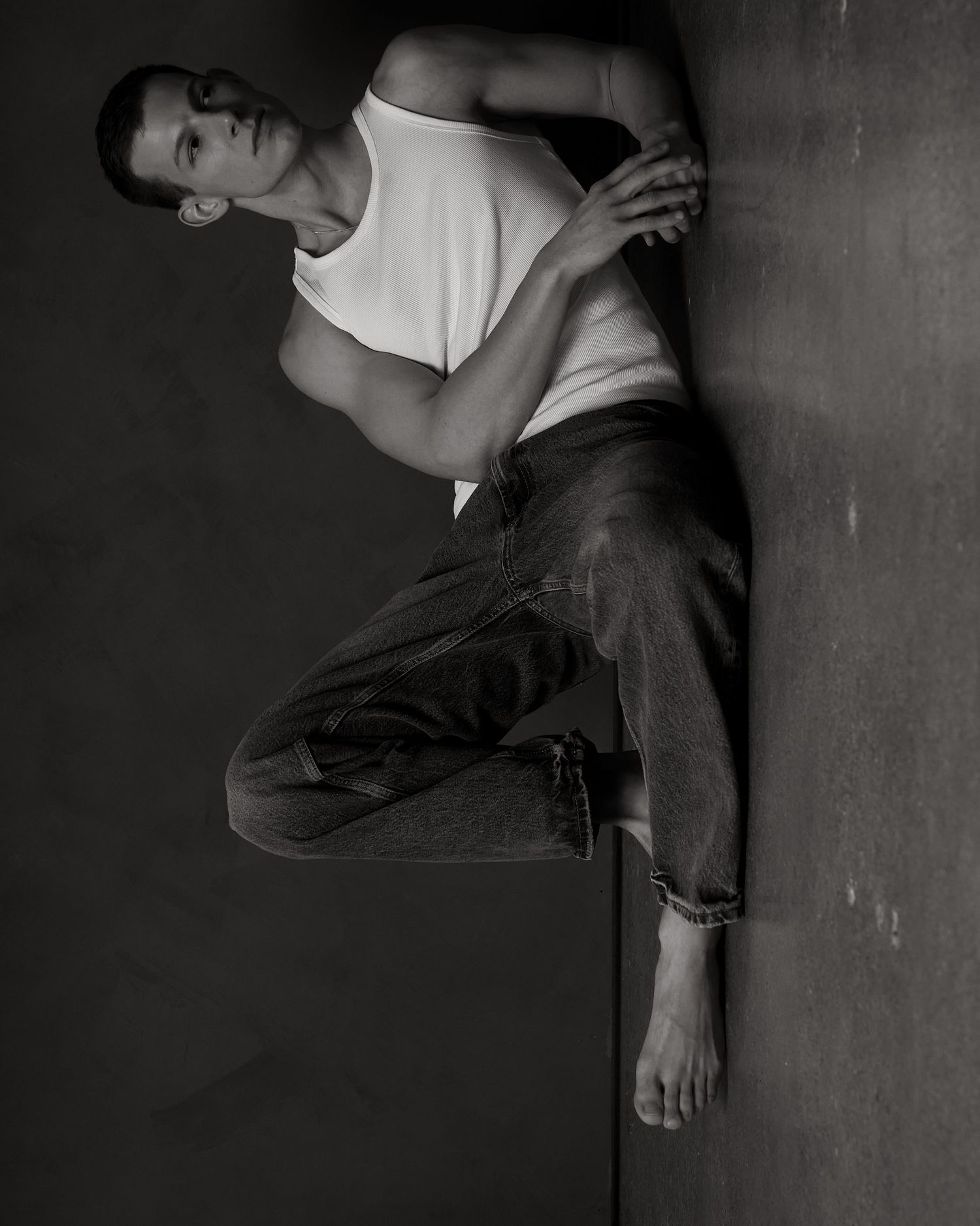 Black and white photo of a man in white tank top and jeans leaning against a dark wall in a dynamic pose.