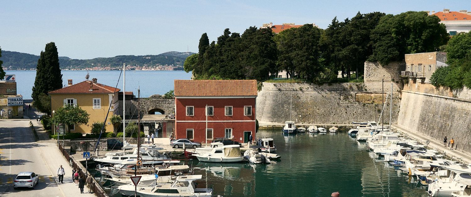 Scenic marina in Zadar, Croatia, with white boats docked near historic stone walls, red buildings, and lush green trees by the Adriatic Sea.