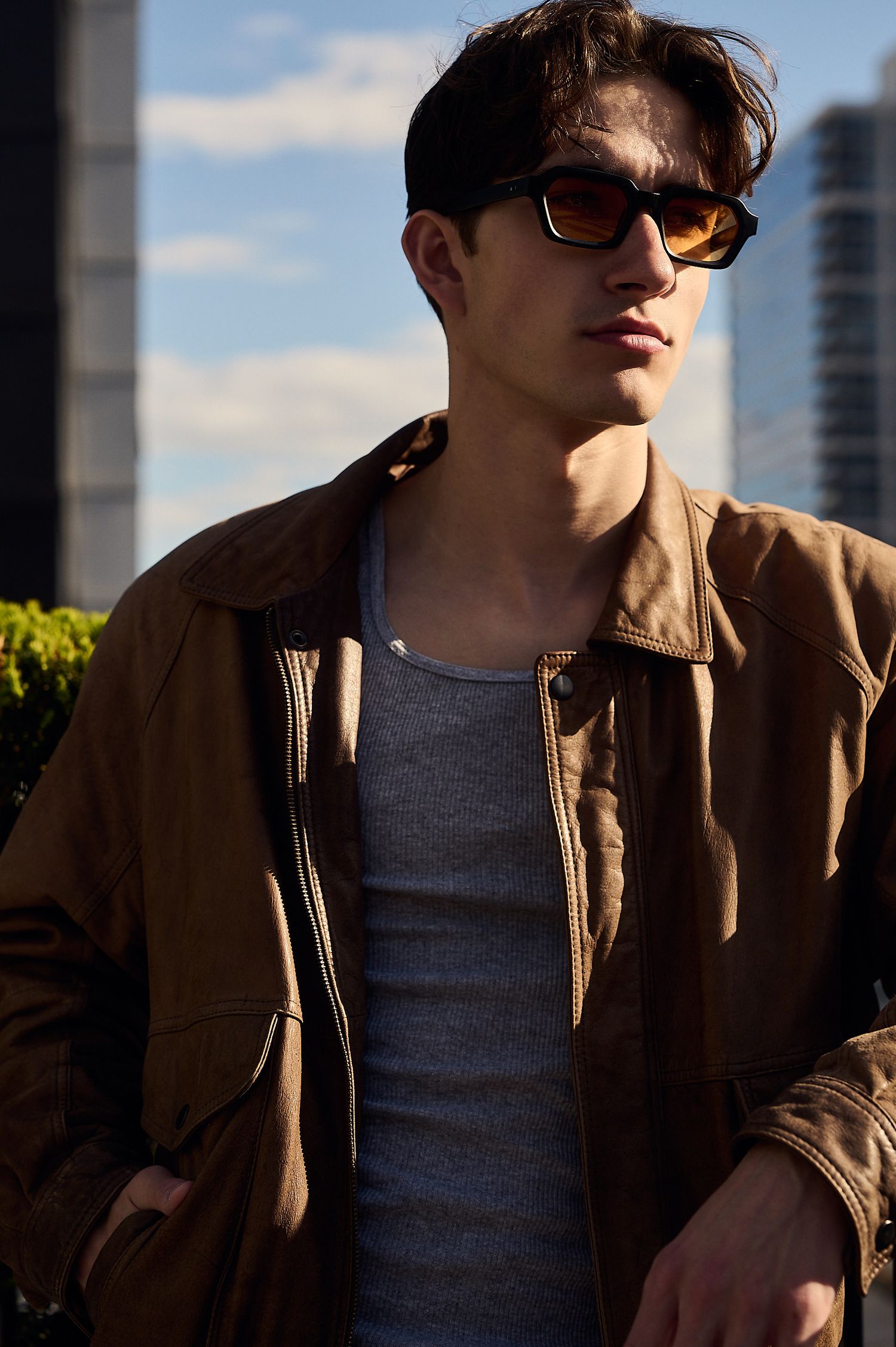 Young man in brown jacket and gray shirt wearing amber sunglasses outdoors with city skyline background.