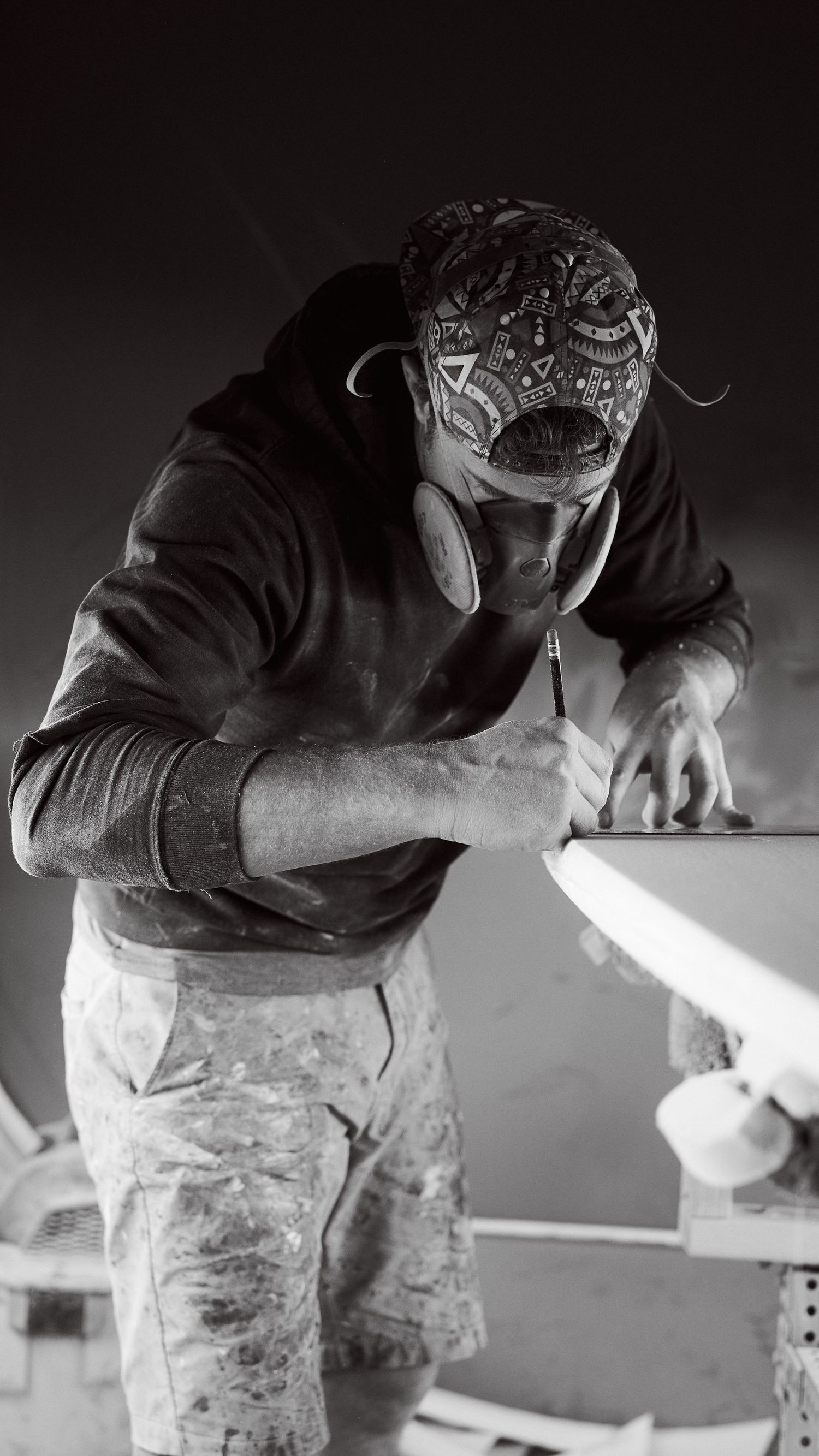 Black and white image of a masked craftsman carefully marking a surfboard in a workshop.