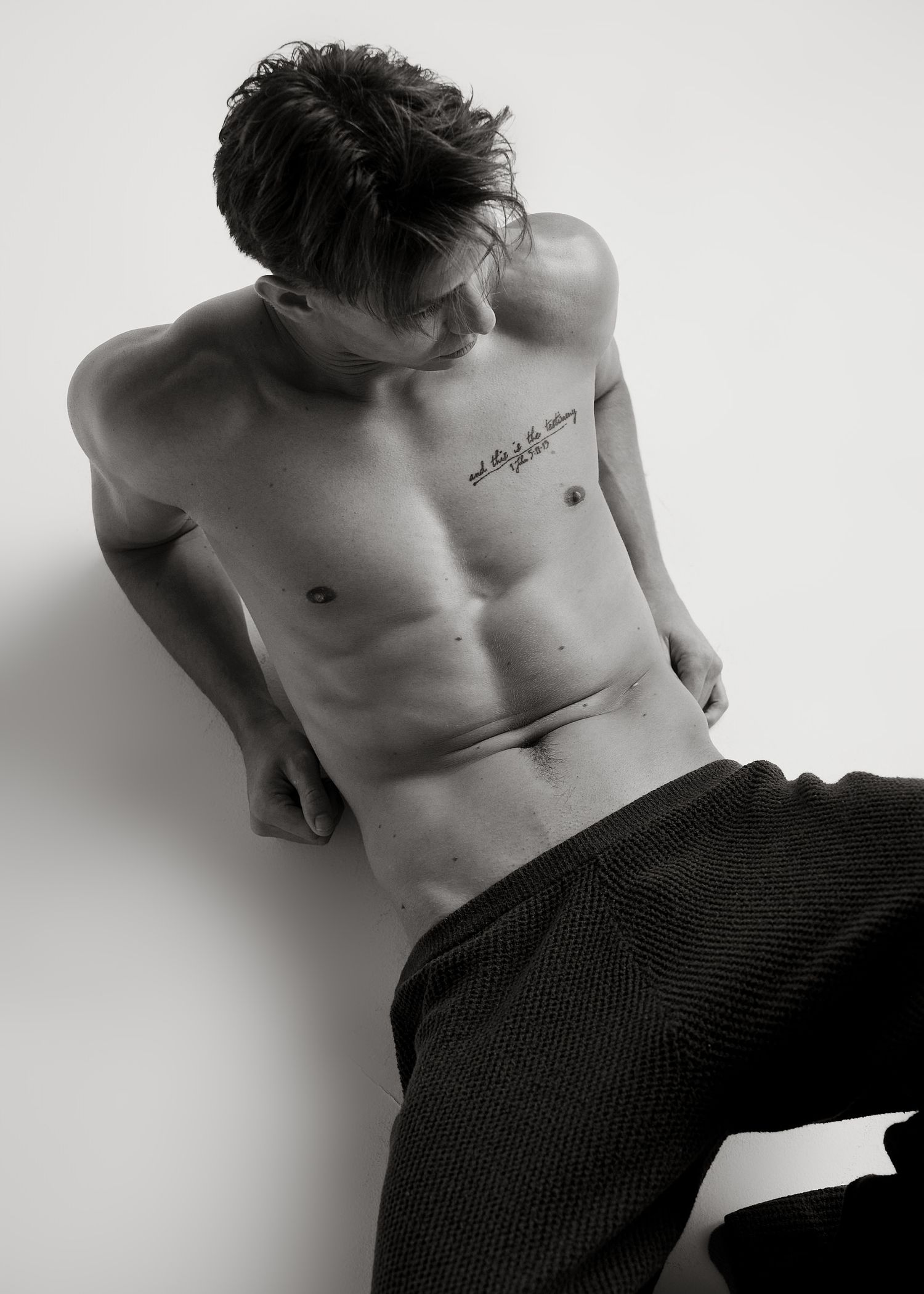 Black and white photo of a shirtless athletic man with chest tattoo wearing dark pants, leaning against a white wall.