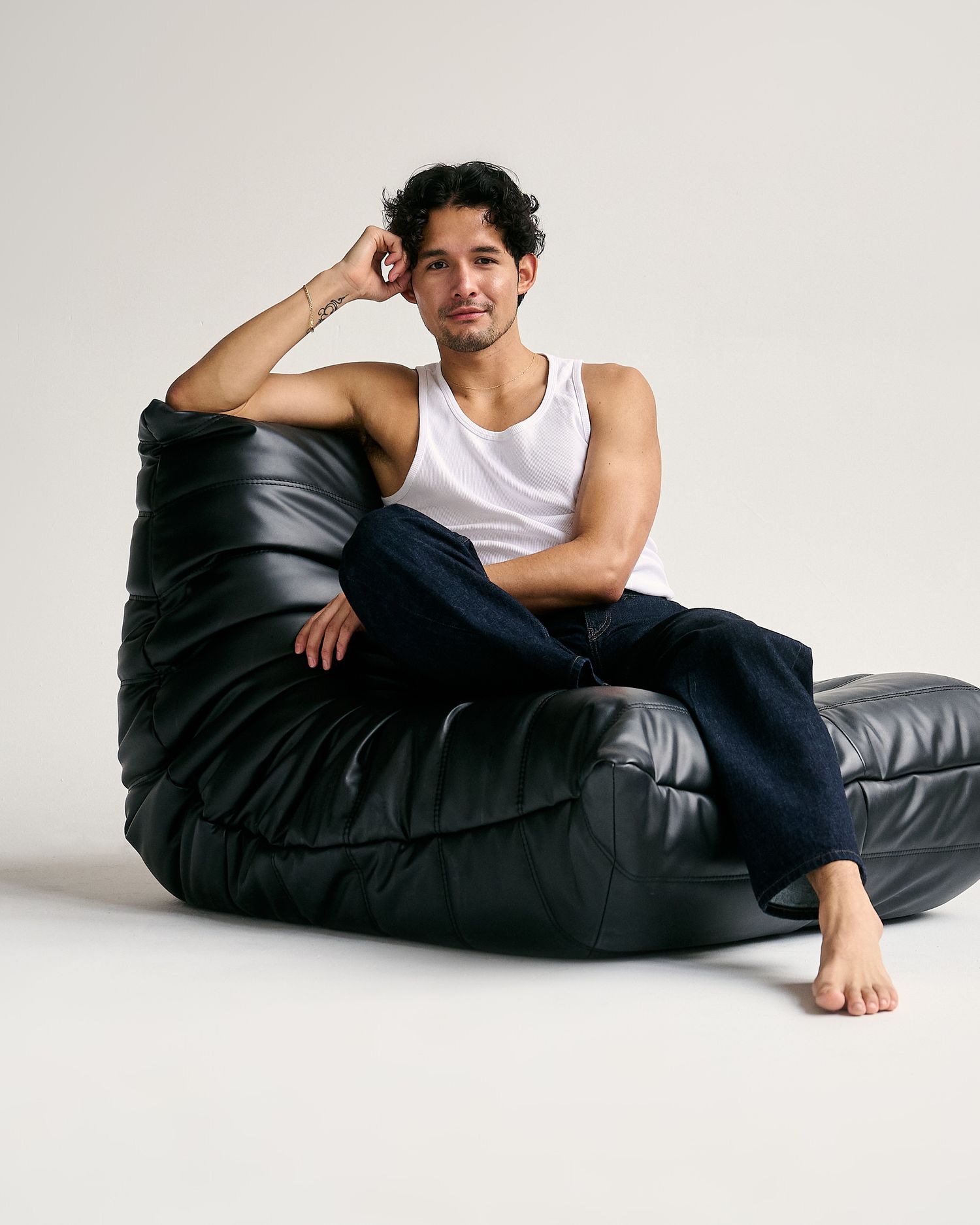Man in white tank top and dark jeans relaxing on a black leather bean bag chair in a studio setting.