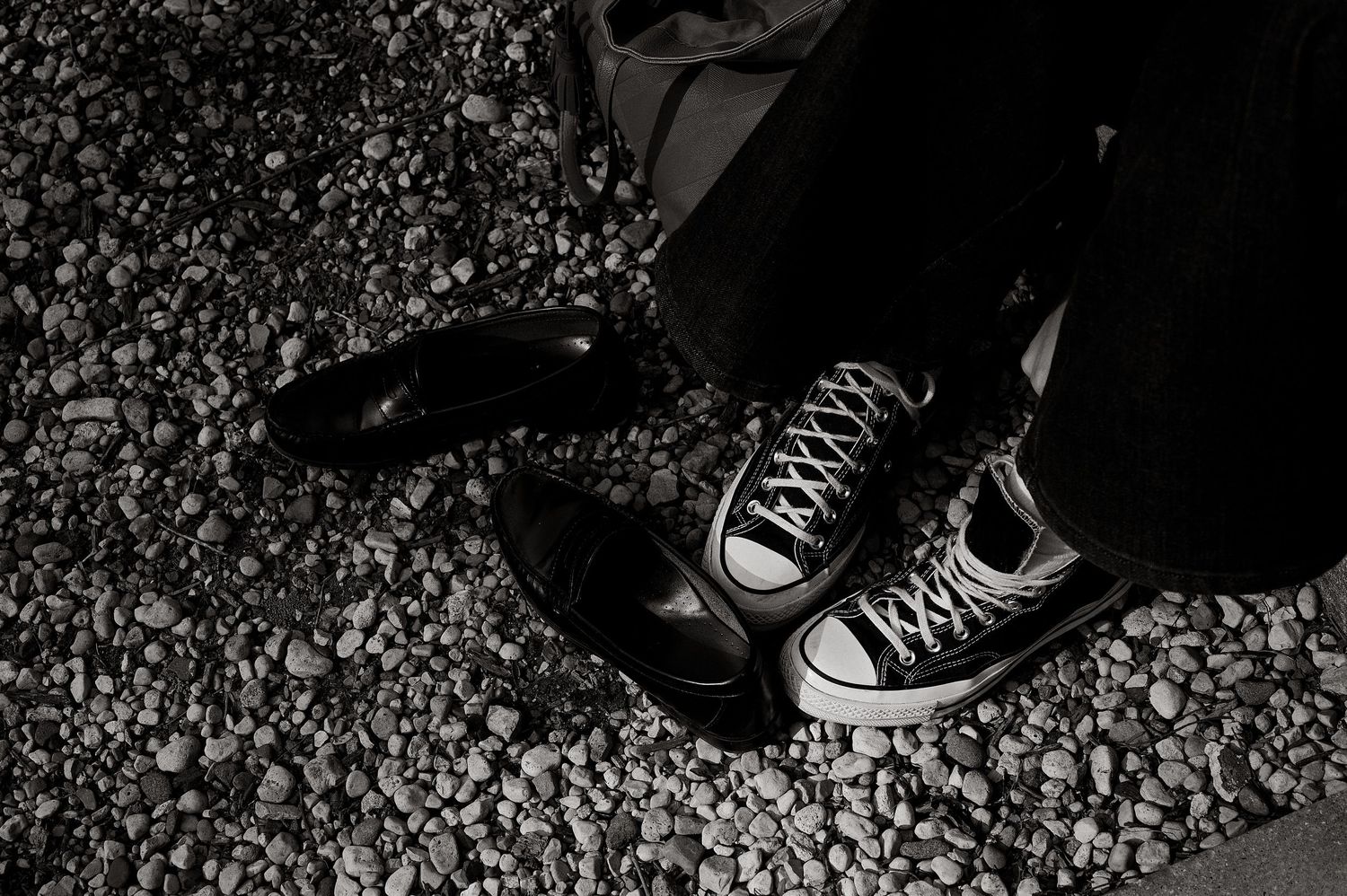 Black and white close-up of two feet wearing mismatched shoes on gravel, one dress shoe and one Converse sneaker.