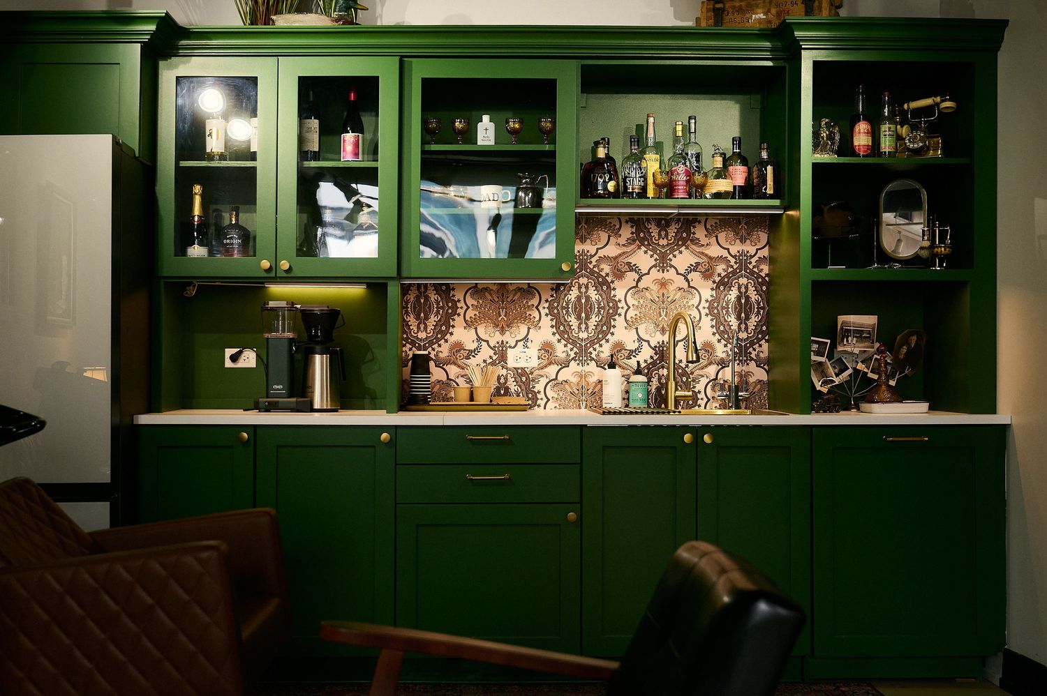 Elegant green built-in bar cabinet with glass doors, decorative wallpaper backsplash, and bottles on display in a stylish home bar.