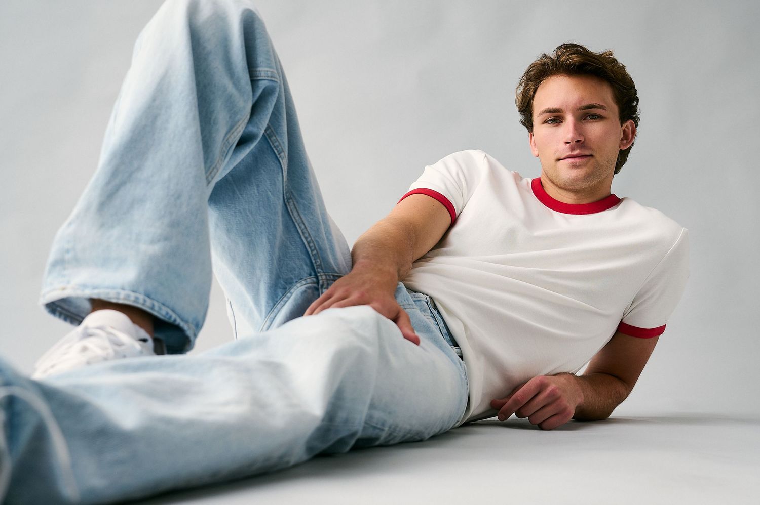 Young man in white ringer tee and light wash jeans sitting casually on white floor in relaxed pose.