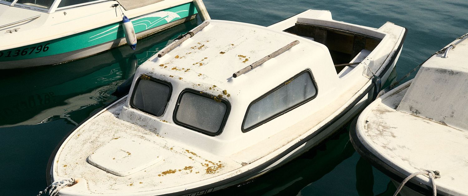 Weathered white motorboat docked at a marina alongside green and red boats on calm turquoise water.