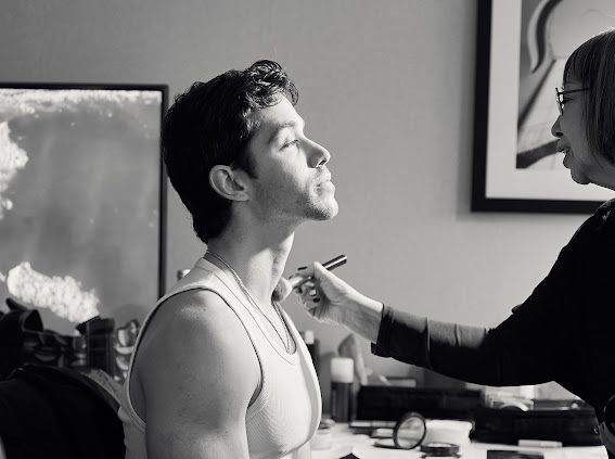 Black and white photo of a man getting makeup applied by a makeup artist in a bright studio setting.