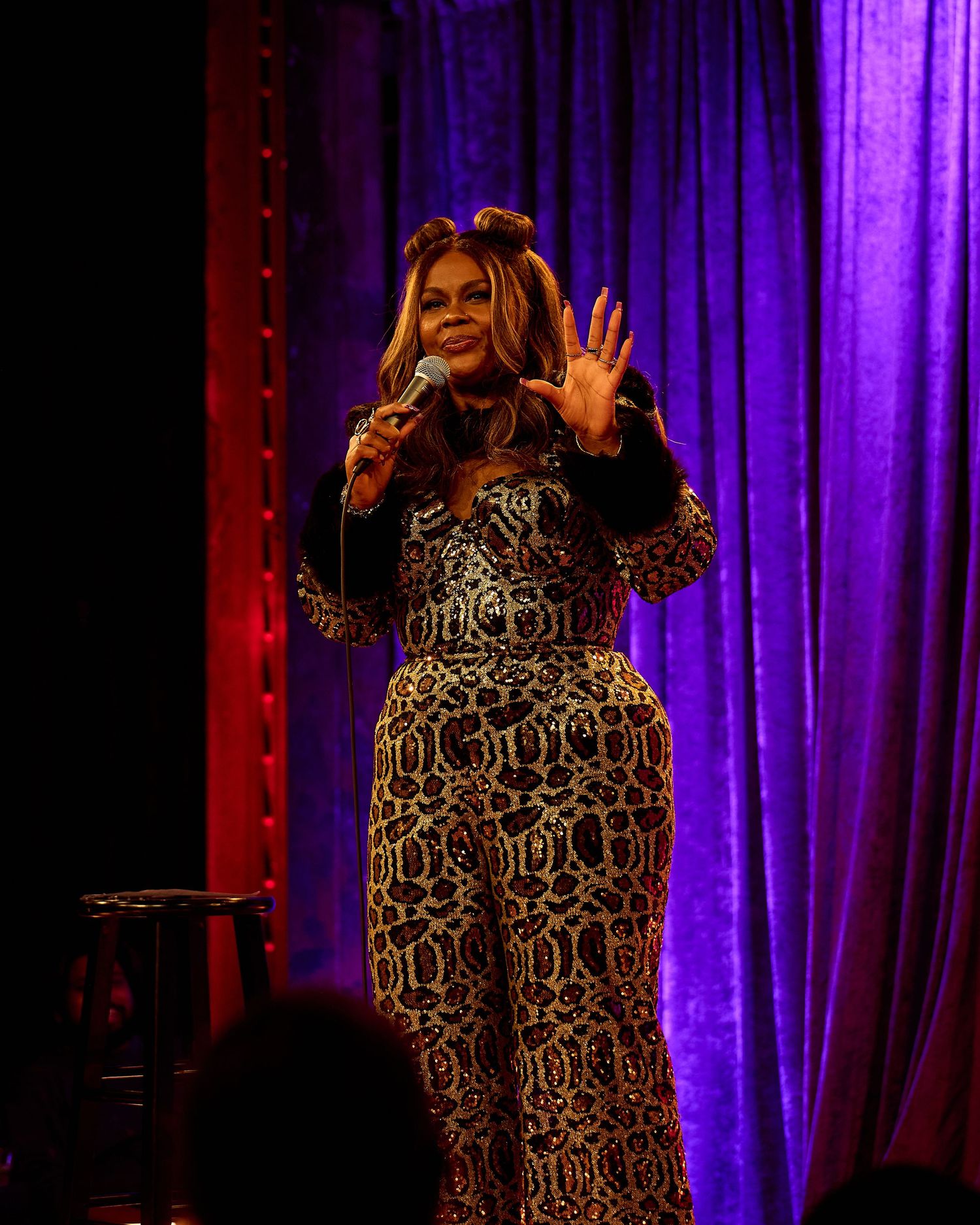 A woman in a gold patterned jumpsuit performs stand-up comedy on a purple-lit stage holding a microphone.