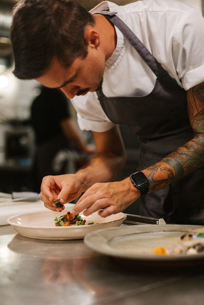 Tattooed chef carefully garnishing a gourmet dish on a white plate in a professional restaurant kitchen.