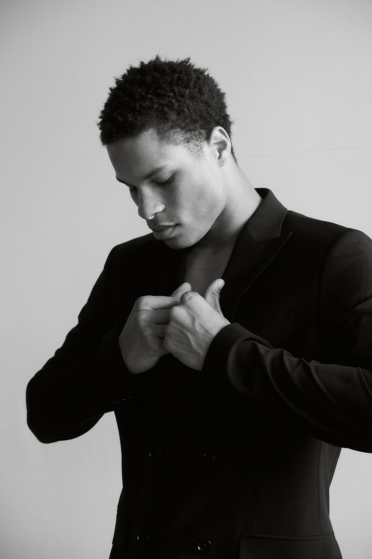 Black and white portrait of a young man in a black blazer looking down with hands clasped at chest.