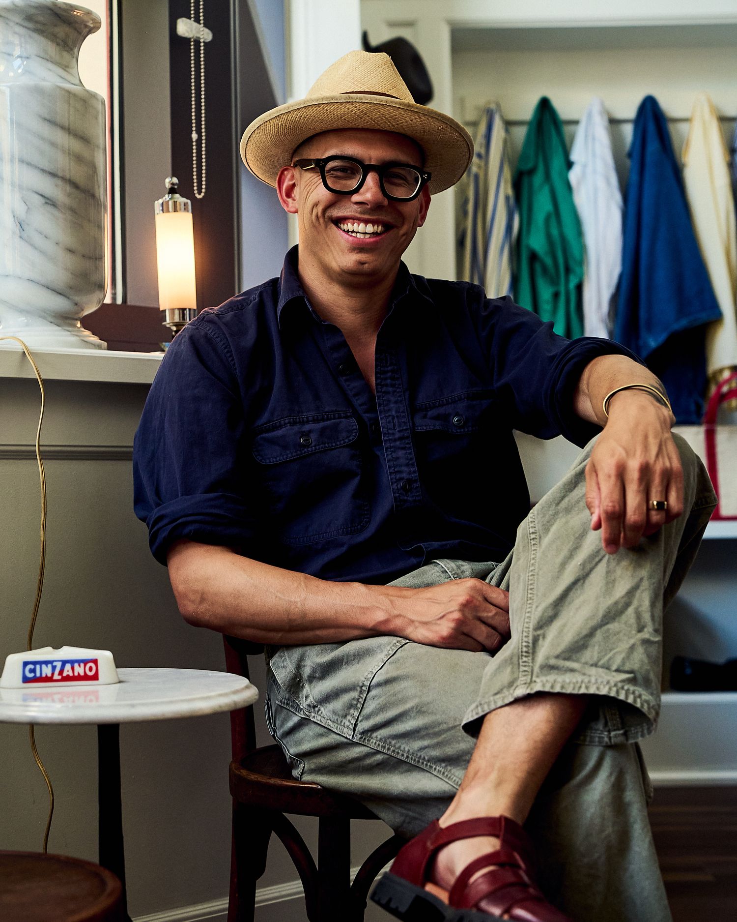 Smiling man in straw hat, dark blue shirt, khaki pants, and red sandals seated in a stylish room with colorful clothing rack.