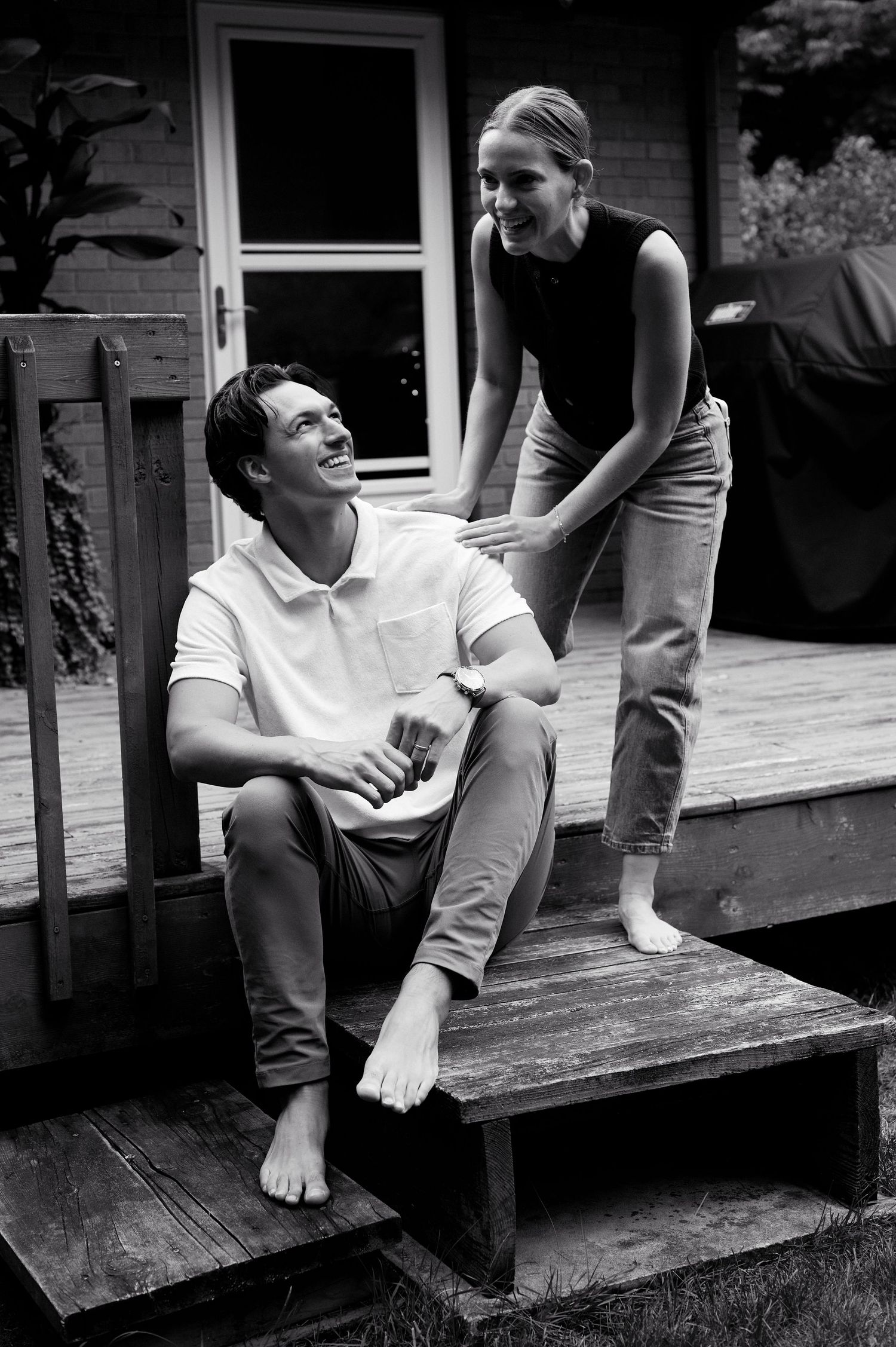 Black and white photo of a couple laughing on wooden porch steps outside a house.