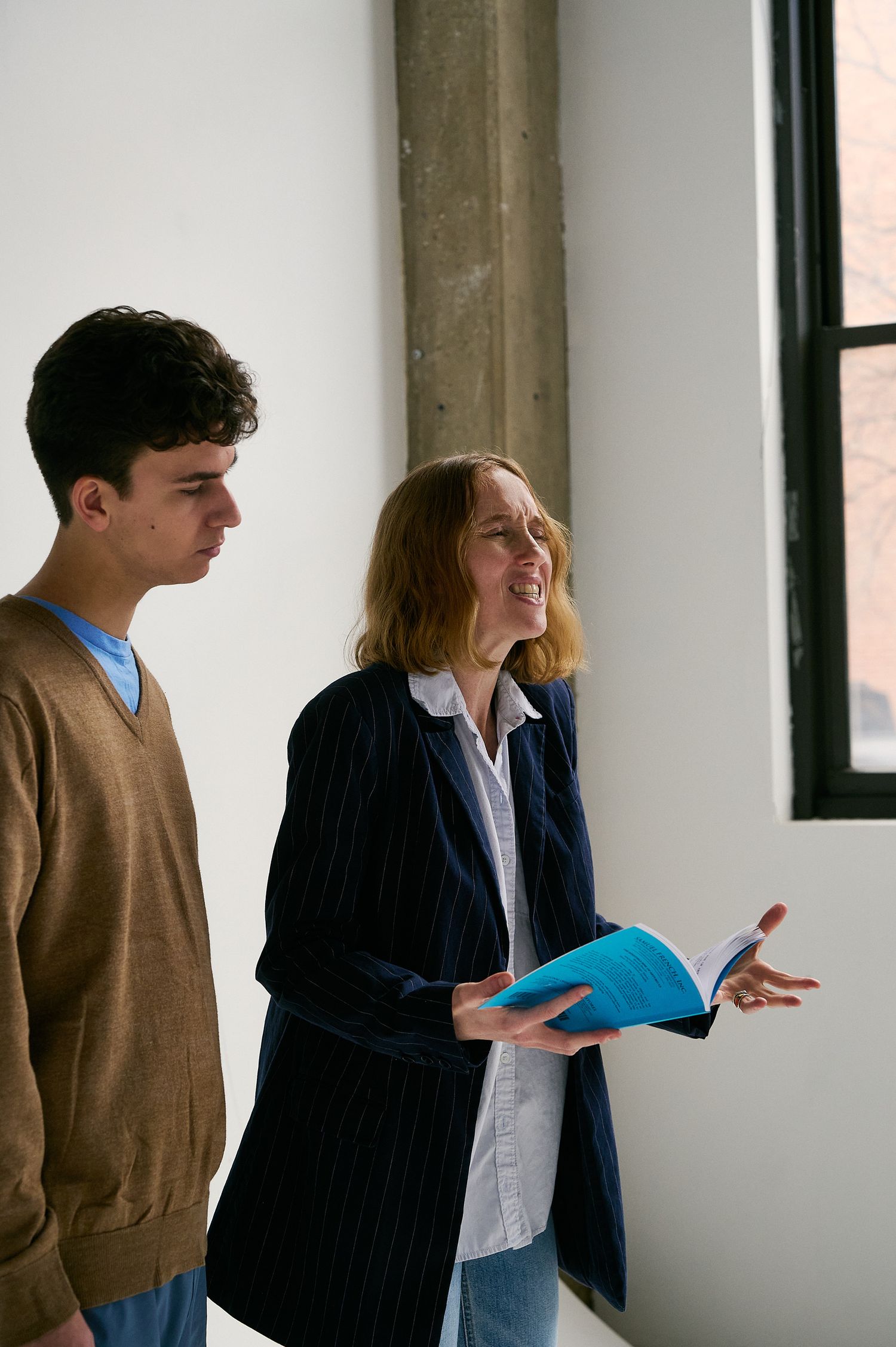 Woman in navy blazer holding a blue book gestures while speaking to a young man indoors.
