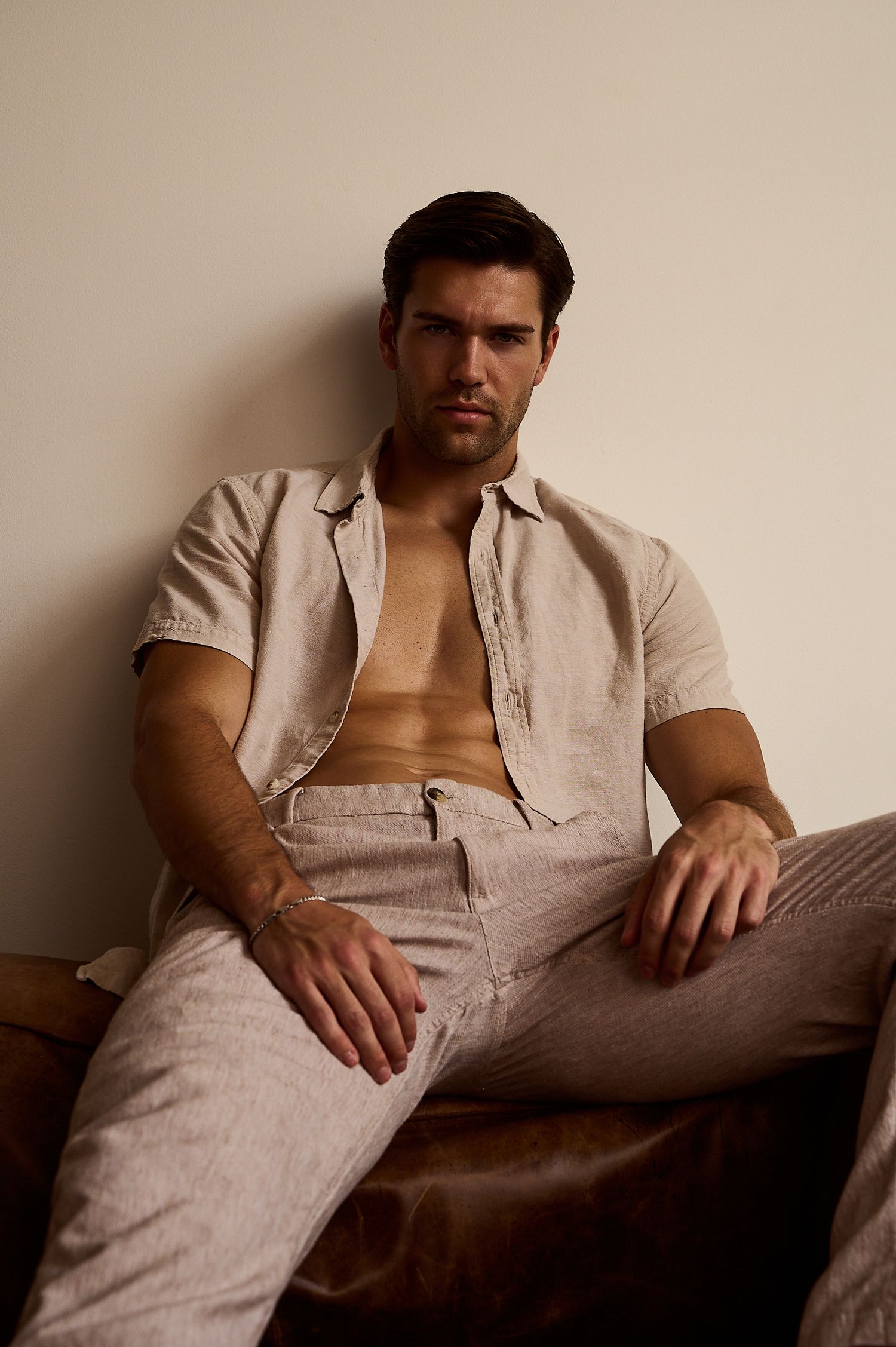 A handsome man in an open linen shirt and matching pants sits relaxed against a beige wall.