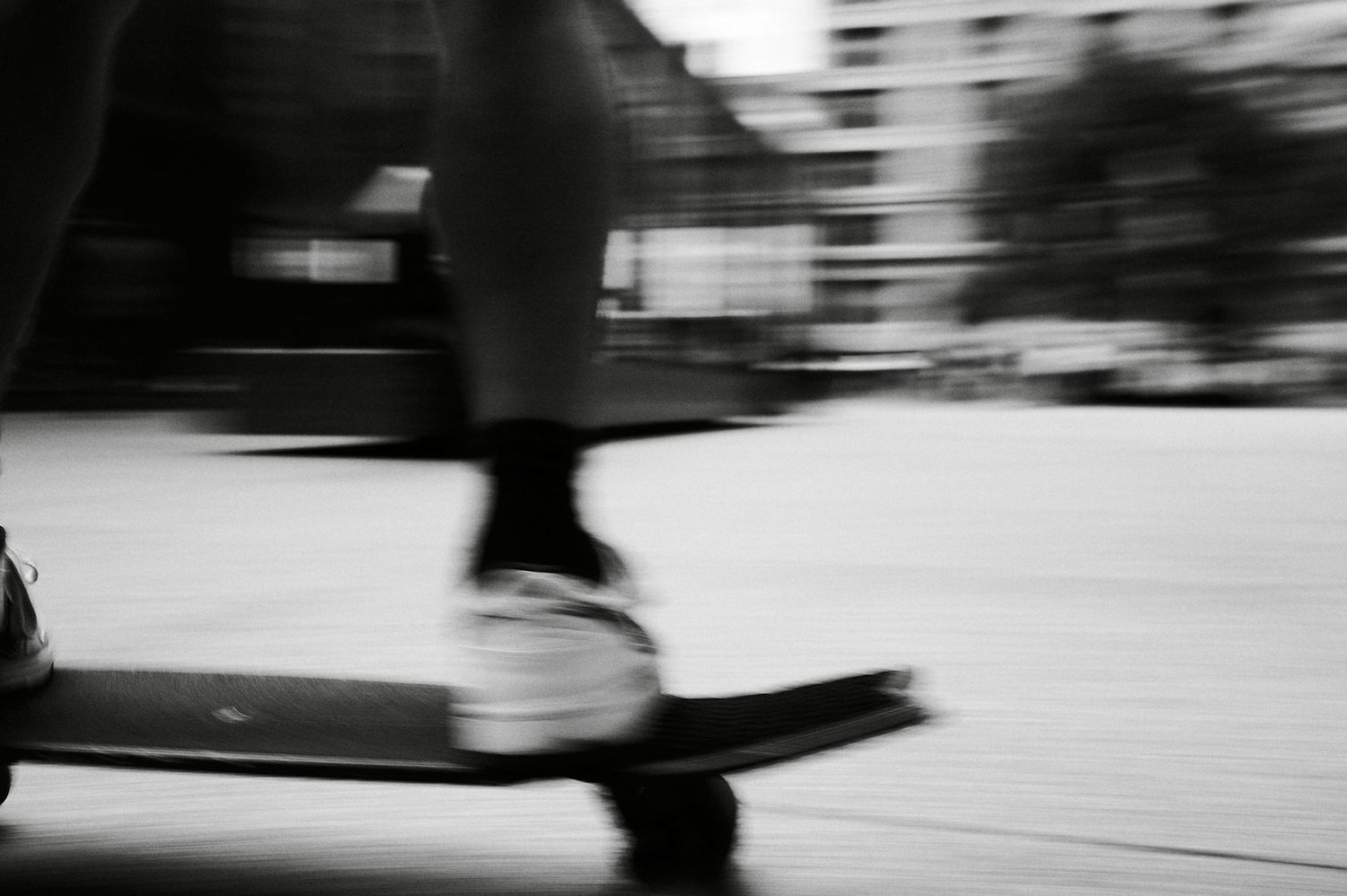 Blurred black and white photo of a person riding a scooter in motion on a city street.