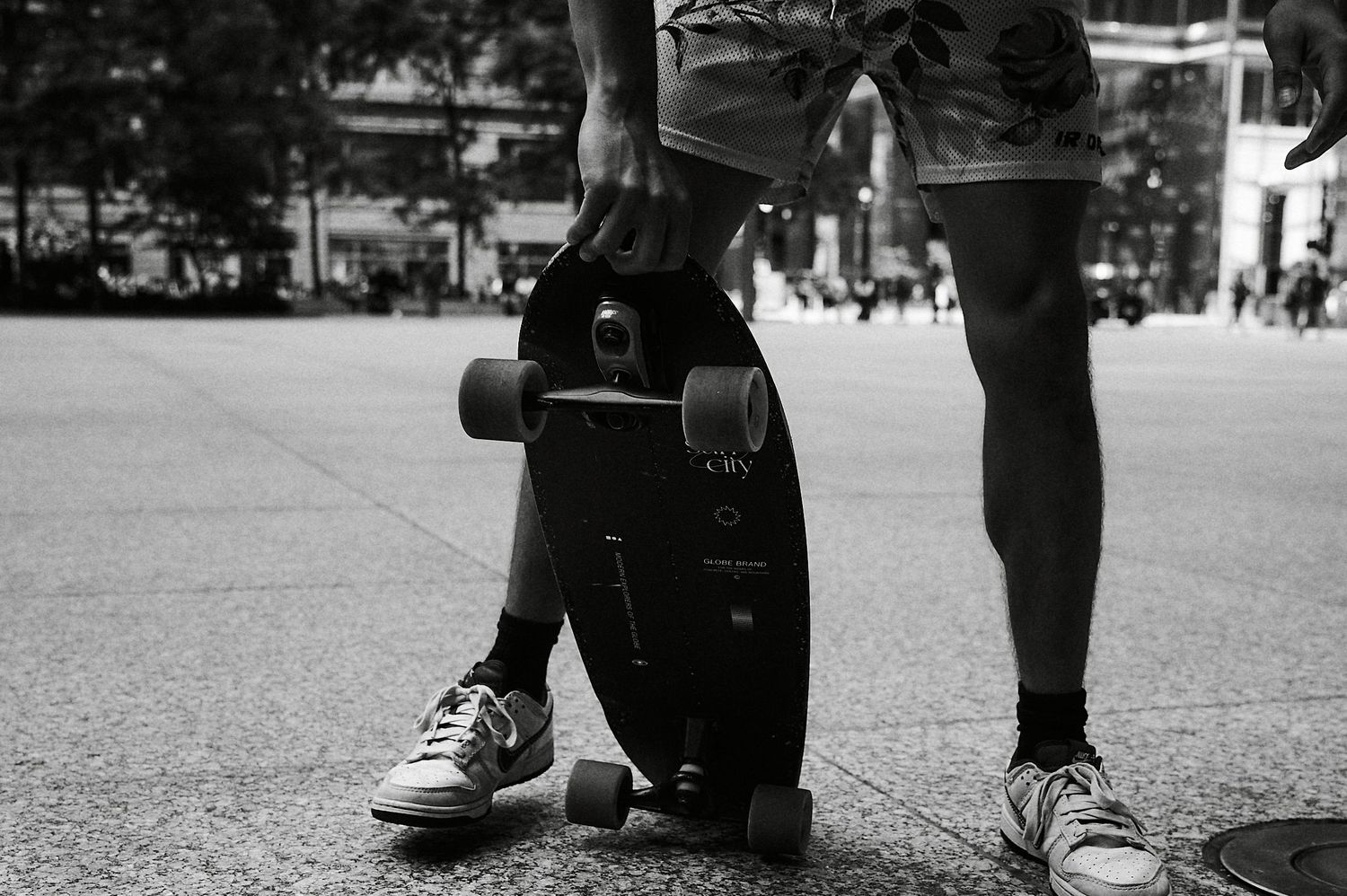 Black and white photo of two people standing with a longboard skateboard on a city plaza pavement.