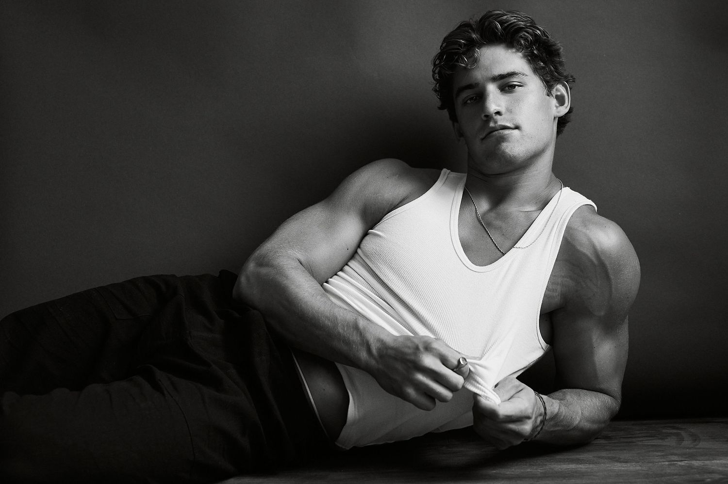Black and white photo of a muscular young man in a white tank top reclining on the floor.