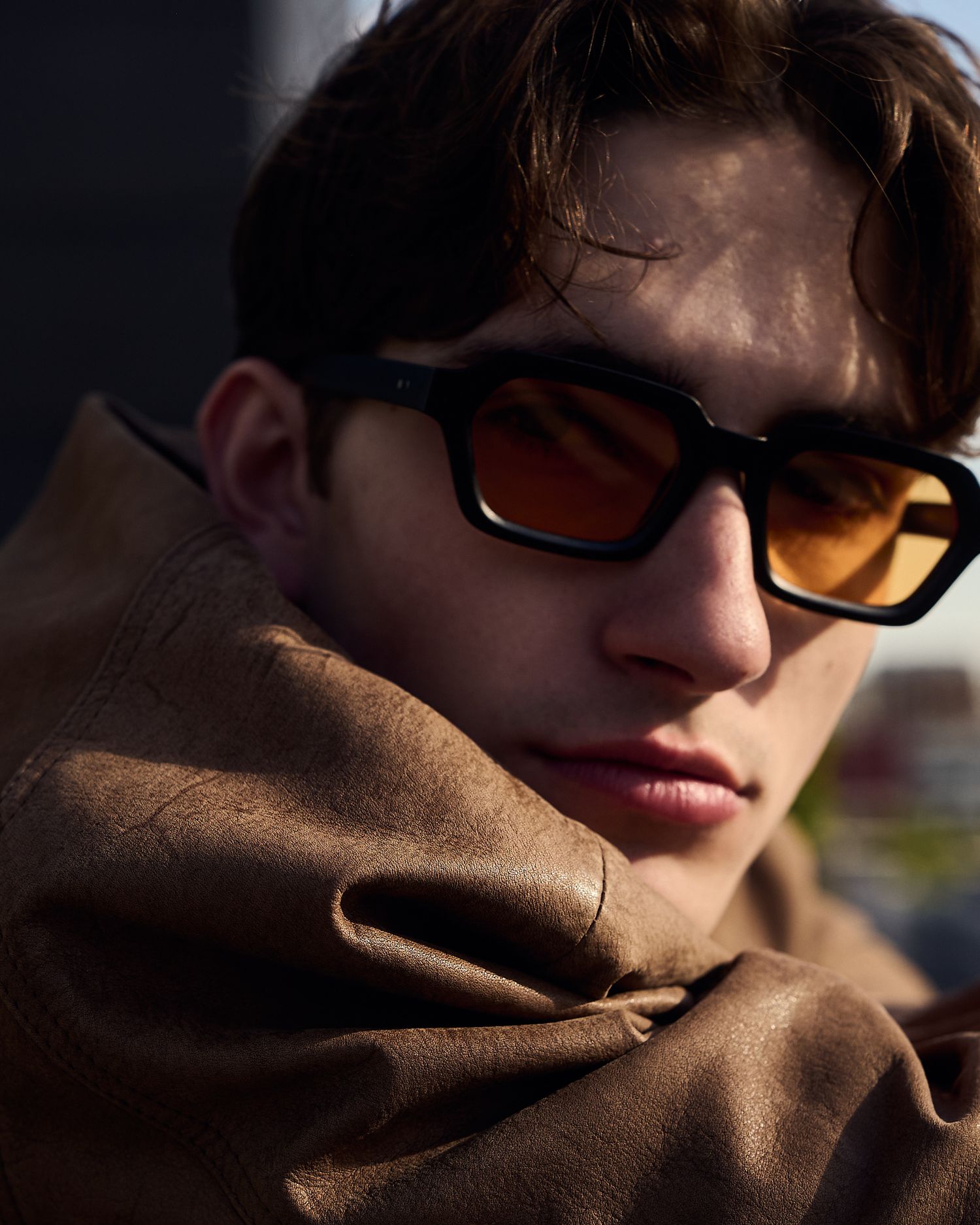 Young man wearing black sunglasses with orange lenses and a brown leather jacket, posed in dramatic sunlight.