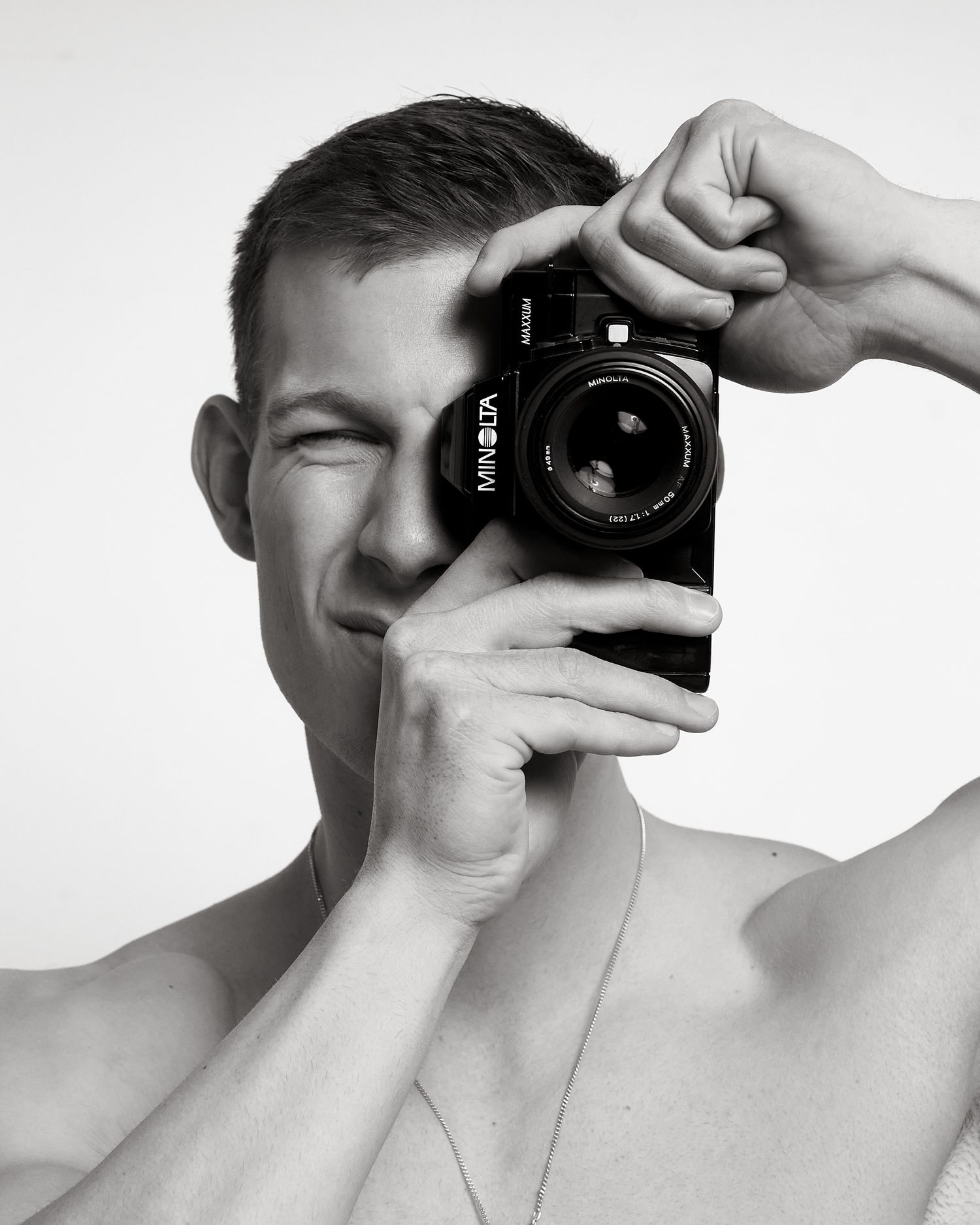 Black and white photo of a young man holding a Minolta film camera up to his face, squinting one eye.