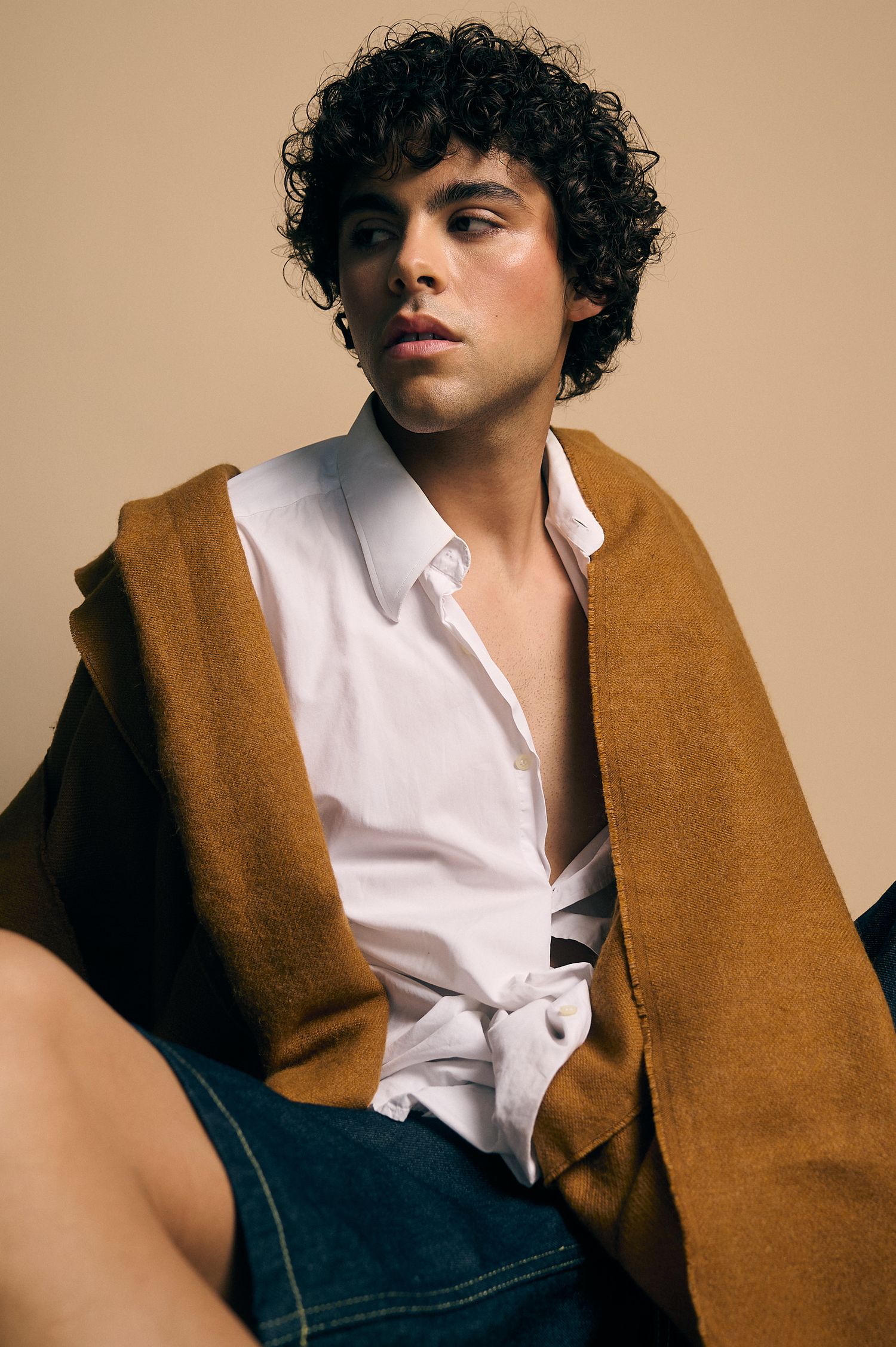 Young man with curly hair wearing an open white shirt and brown draped jacket, seated against a beige background.