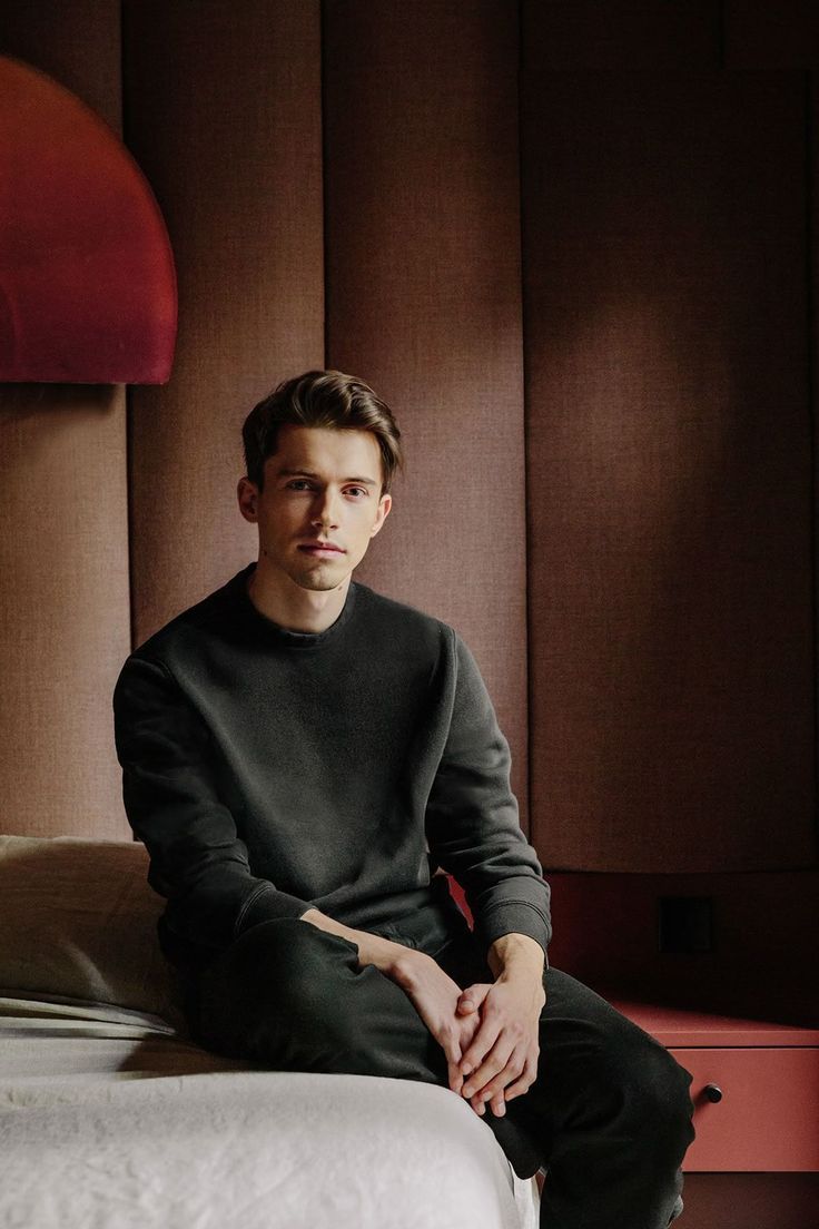 Young man in dark green sweatshirt sitting on bed in modern room with brown paneled walls.
