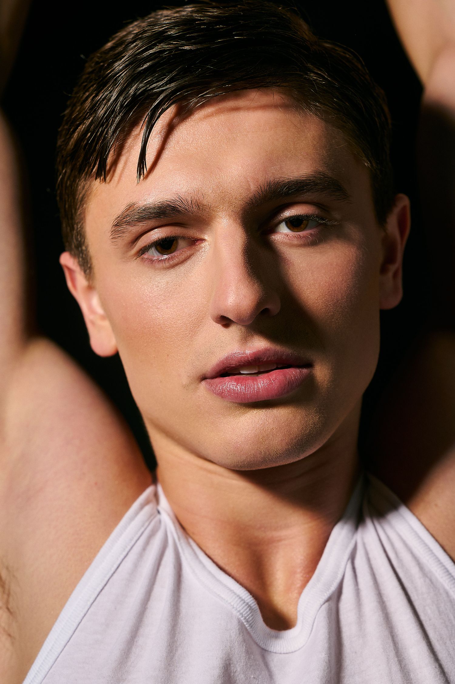 Close-up portrait of young male model with wet hair in white tank top under dramatic lighting.