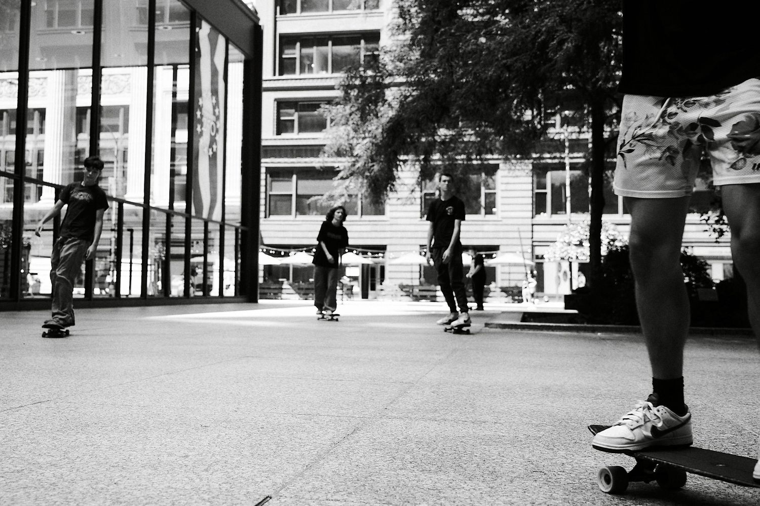 Black and white photo of skateboarders skating in an urban plaza with buildings in the background.