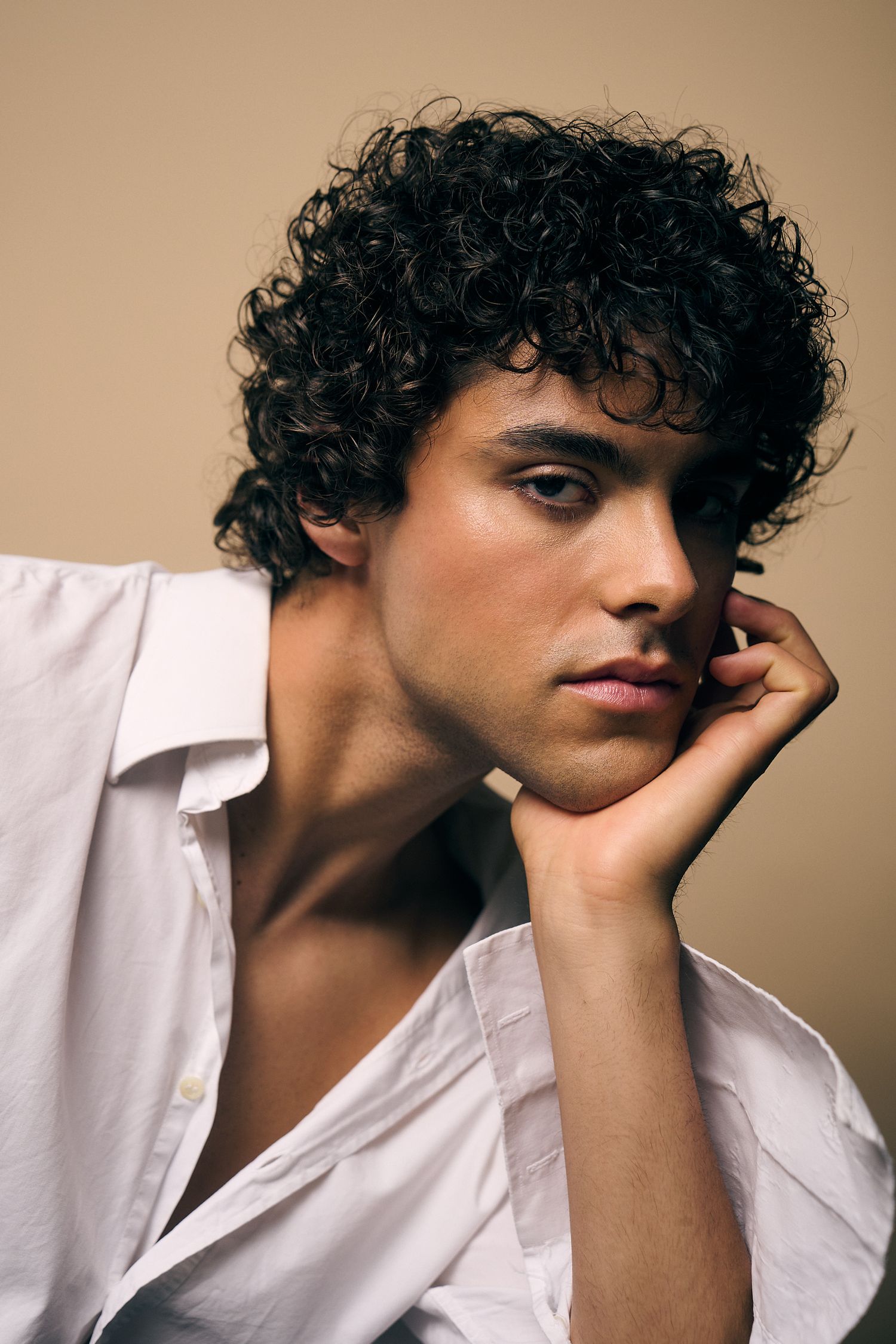 Young man with dark curly hair in white shirt resting chin on hand, studio portrait with neutral background.
